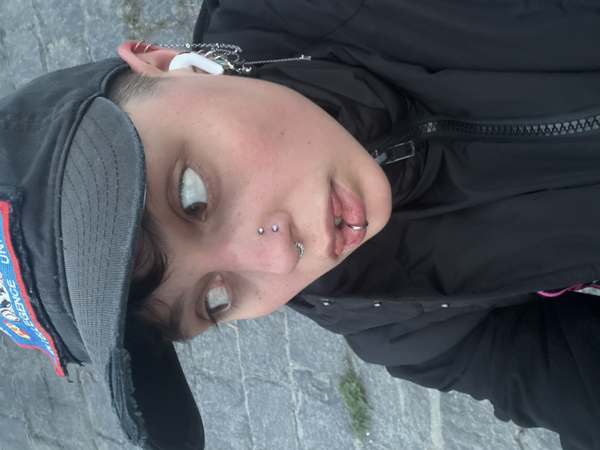 The image displays a close-up portrait of a young person, captured outdoors at a low angle and rotated 90 degrees clockwise. The subject has short hair and is wearing a dark baseball cap with a blue and red patch on the side, featuring partial white text that includes "UNIVERSITY" and "UNI". They are also dressed in a black puffer jacket with a visible zipper down the front.
The person's head is tilted back, and their eyes are rolled upwards, showing a significant amount of white sclera, with their brown irises visible at the bottom. Their mouth is slightly open, revealing a silver ring piercing on their lower lip. They have multiple facial piercings, including two studs on the bridge of their nose and a segmented ring in their left nostril. Their left ear is adorned with several piercings, including a hoop at the top helix, and a white earbud is visible, with a chain draped from it connecting to other ear jewelry.
The background consists of grey paving stones, characteristic of urban settings, with sparse green vegetation growing in the cracks. The lighting suggests it is daytime. The overall pose appears stylized. The setting is in Saint Petersburg, Russia.