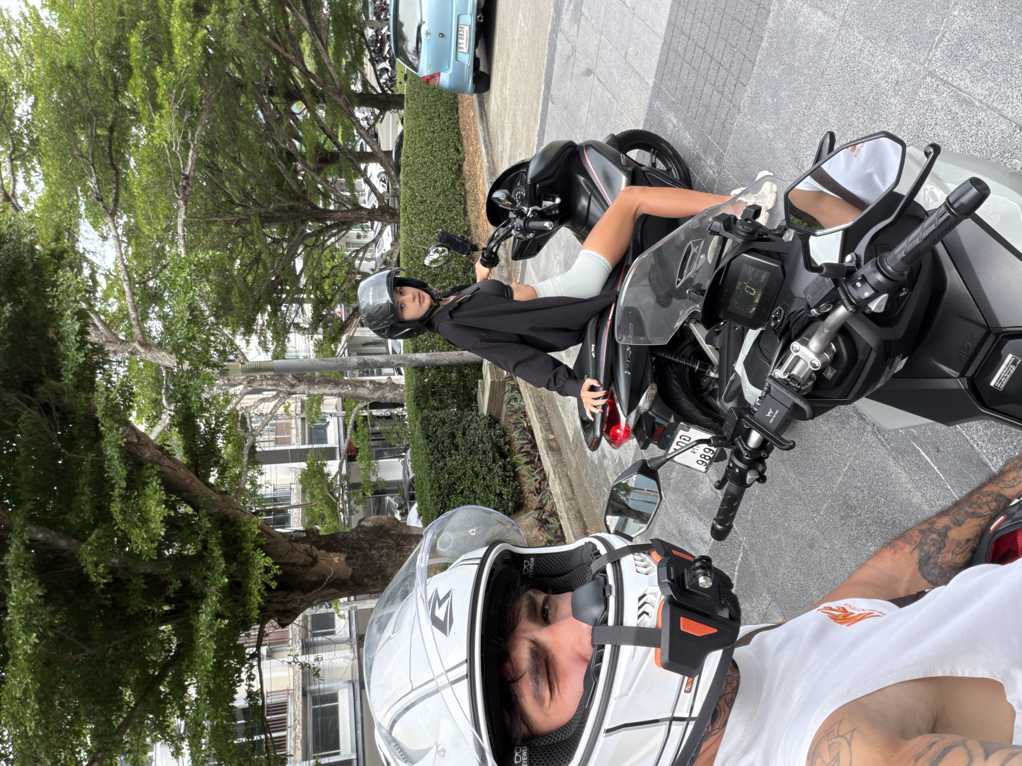 Two individuals are present in a selfie-style photograph. The photographer, wearing a white helmet, white t-shirt, and displaying arm tattoos, is positioned at the bottom.
To the rear of the photographer, a woman sits astride a black and white motorbike. She wears a black helmet and a black hoodie.
Two motorbikes are visible in the scene. A blue car is parked in the background. A leafy tree is also evident.
The location is Chalong, Thailand.