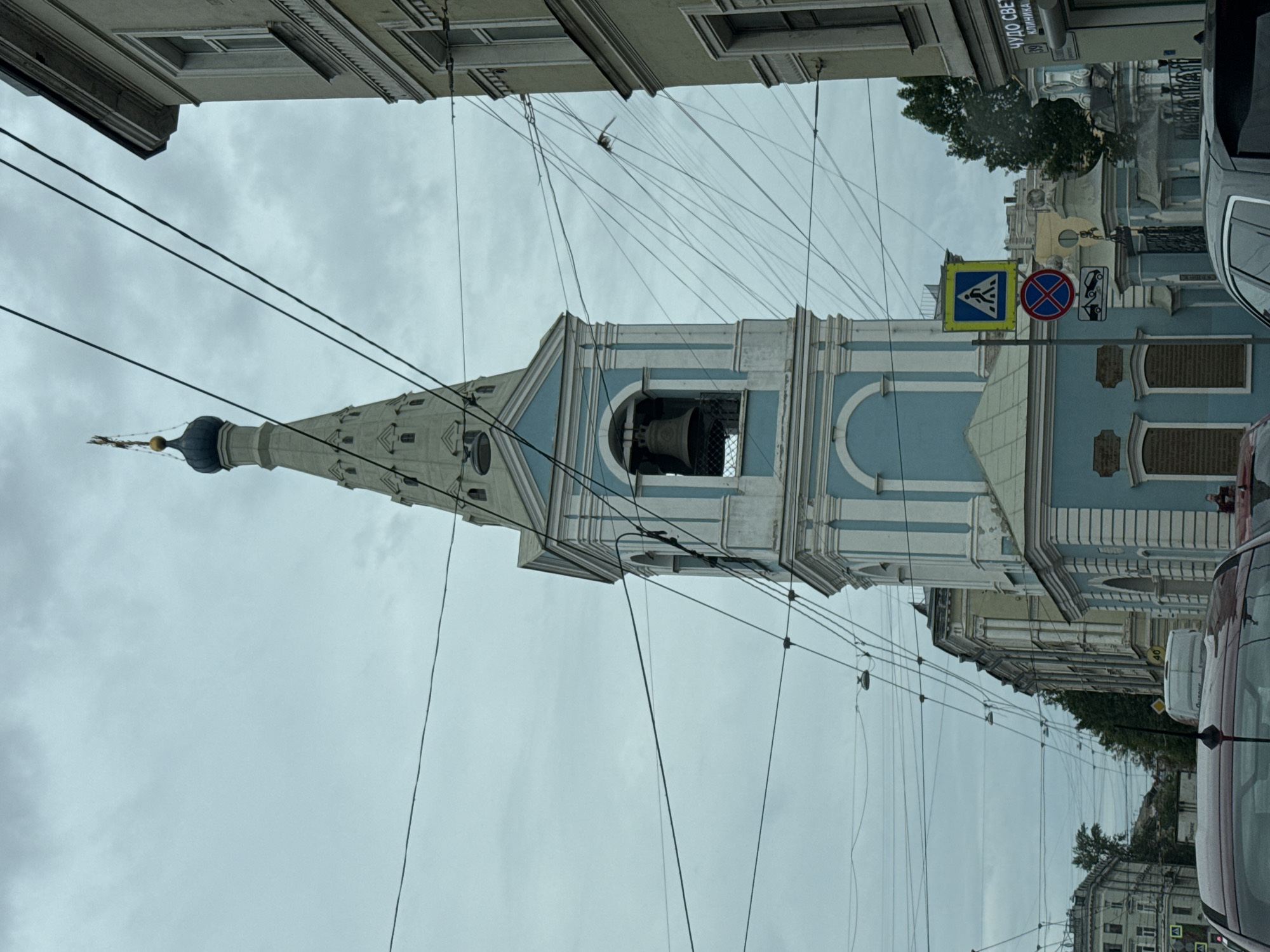 The image captures a street view in Saint Petersburg, Russia, focusing on the bell tower of the Church of the Smolensk Icon of the Mother of God. The bell tower is tall and slender, painted light blue and white, with a gilded cross atop its dark blue dome. A large bell is visible within an arched opening in the tower. The sky is overcast, with a dense network of overhead wires crisscrossing the frame, typical of an urban environment. To the right of the bell tower, the corner of a light yellow building is visible, featuring classical architectural details. In the foreground, the hoods of two cars are visible, suggesting the photograph was taken from a vehicle. Further down the street, more buildings and trees can be seen. Several street signs are also present, including a pedestrian crossing sign and a prohibition sign. The overall impression is of an ordinary day in a historic city, with the church spire being the dominant architectural element.