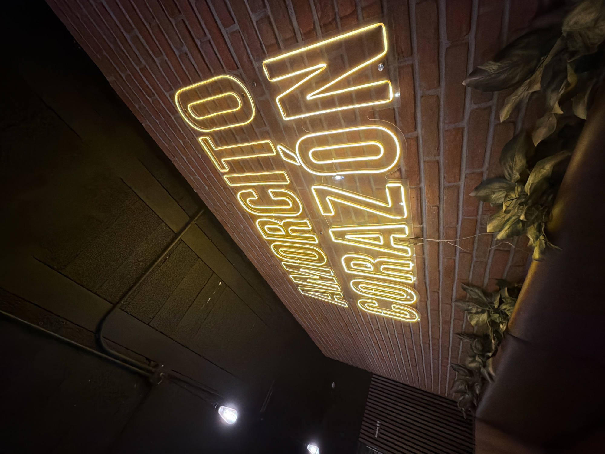 The image depicts the interior of a restaurant or bar in an unknown city. The focal point is a neon sign mounted on a brick wall, reading "NO CITÓ AMORCITO CORAZÓN." The sign is illuminated in a warm yellow light. To the right of the sign, there are green leafy plants, likely part of the decor. The left side of the image is dominated by a dark wall, possibly painted black, with some visible pipes or conduits running along it. There are a few dimly lit spotlights on the dark wall. The overall ambiance is somewhat dim, suggesting a cozy or intimate setting. A partial view of dark furniture, possibly a booth or seating, is seen in the lower right corner. The restaurant has a contemporary style.