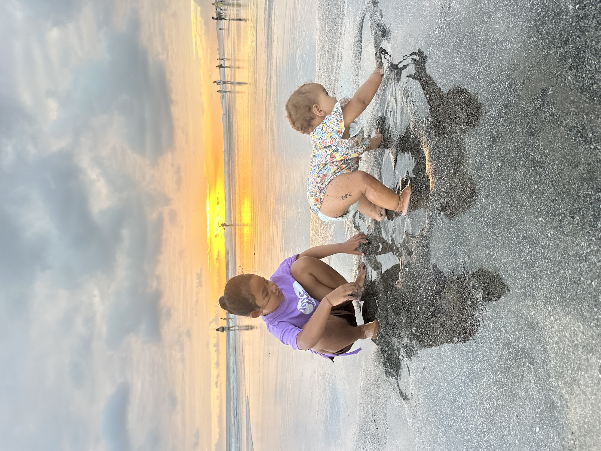 In the city of North Kuta, Indonesia, at sunset on a beach, two young girls are playing in the wet sand near the ocean. The older girl, wearing a purple t-shirt and brown shorts, is crouched down and appears to be digging in the sand with her hands. The younger child, a baby wearing a floral-patterned romper, is on her hands and knees, also playing with the wet sand. The sky is filled with dramatic clouds, with a bright orange and yellow sunset visible on the horizon over the ocean. Several other people are visible in the distance, walking along the beach. The wet sand reflects the light of the sunset, creating a glossy surface.