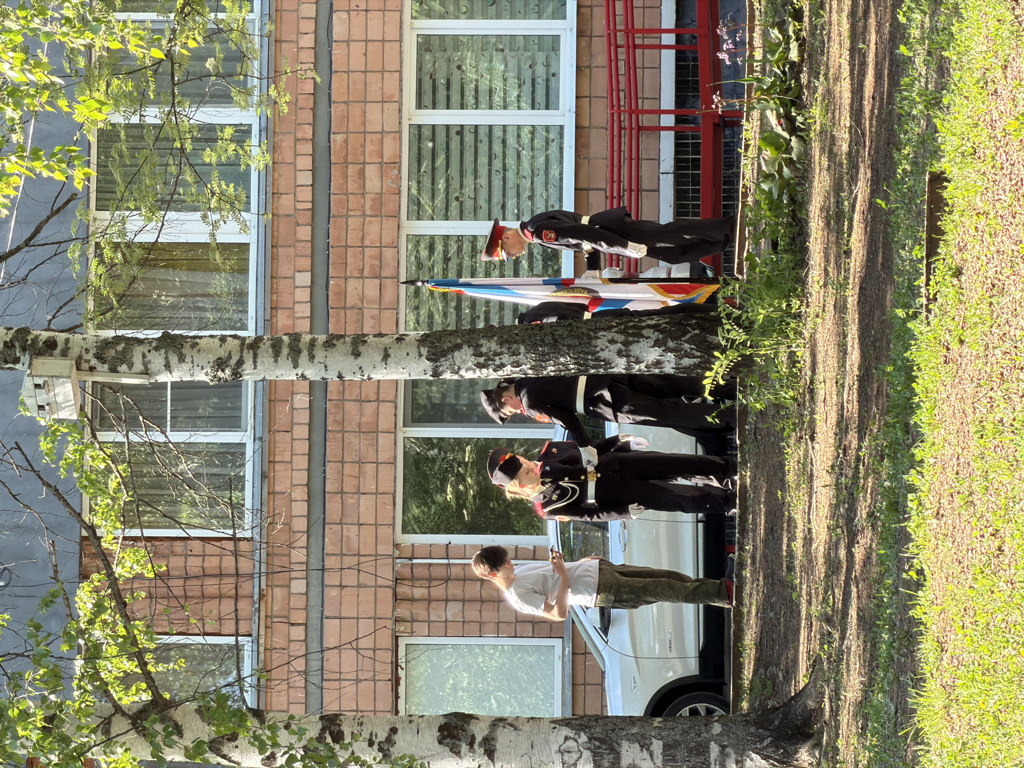 The image captures an outdoor scene in Забегалово, Russia, featuring three individuals in what appear to be formal military or cadet uniforms standing at attention behind a flag. A civilian in a white shirt and khaki pants is standing in front of them. They are standing in front of a brick building with windows. A birch tree is visible in the foreground. To the side is a white SUV parked on a paved area. Grass is on the far right. The sun is shining, casting shadows on the ground.