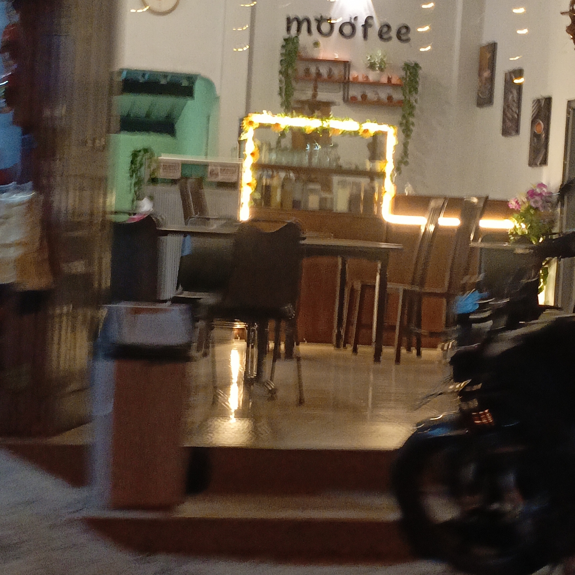 The image depicts the interior of a cafe named "mooffee" in Salatiga, Indonesia. The cafe features a modern, dimly lit ambiance with warm lighting, likely from neon or LED strips framing a display area and overhead fixtures. Visible elements include wooden tables and chairs, shelves stocked with items, and decorative plants hanging from the ceiling. In the foreground, a portion of a black motorcycle is visible, suggesting it is parked in or near the cafe. The overall scene suggests a casual dining or coffee shop environment.