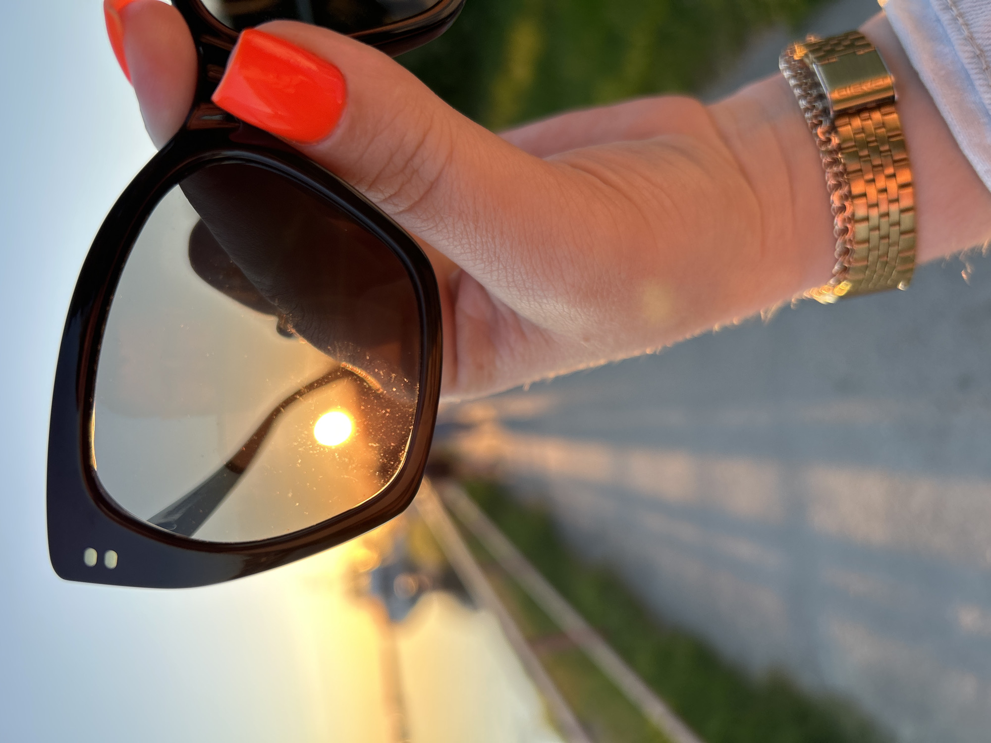 The image presents a first-person perspective, focused on a light-skinned hand holding a pair of dark-framed cat-eye sunglasses. The fingernails are painted a vibrant neon orange/red, uniformly short and rounded. A gold-toned, multi-link metal watch with a clasp is visible on the wrist. The sunglasses feature a dark, likely black or deep brown, frame with two small, light-colored circular studs on the upper left corner. The lenses are reflective, displaying a prominent, bright, circular orange-yellow sun at a low angle, indicative of sunset. Fine specks or dust are visible on the surface of the lens.
The background shows an outdoor environment during sunset, characterized by a gradient sky transitioning from light blue at the top to warm orange and yellow near the horizon. To the left, a calm body of water, potentially a river or bay, reflects the ambient light. A paved pathway or promenade extends into the distance, bordered on the left by a light-colored, possibly white or wooden, railing or fence that separates the path from the water. Long shadows are cast across the pathway, consistent with the low sun angle. Indistinct, blurred shapes in the middle distance suggest boats or structures on the water, and faintly visible human figures are discernible walking on the distant path.
Based solely on the visual information within the image, no specific landmarks, architectural styles, or unique geographical features are present to definitively confirm the location as Tallinn, Estonia. The scene depicts a generic waterfront promenade at sunset.