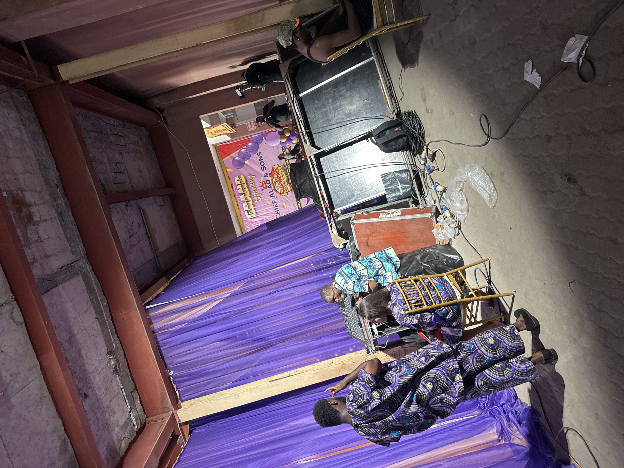 The scene is a room in Lagos, Nigeria, prepared for an event. The room has exposed ceiling beams and unfinished walls. A purple drape is in place as a backdrop, behind which is a banner that says "Supreme Chief Alao & Sons Opening". Balloons decorate the scene. A sound system with a sound mixer is set up. Three people are present: one stands in front of the mixer, and two others sit on chairs nearby. Cables are on the floor.