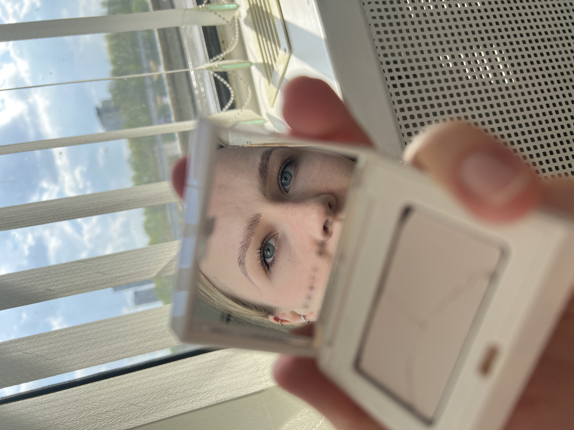The image depicts a person holding a white, square compact mirror. The reflection in the mirror shows a partial view of a face, including light blue eyes, light eyebrows, and a portion of the nose and cheek. An earring is visible in the reflected ear.
The background includes a window with closed horizontal blinds. Outside the window, there is a blurred view of buildings and trees under a cloudy sky.
The location of the scene is specified as Khimki, Russia, although there are no specific visual cues within the image to directly confirm this location. However, the presence of urban buildings and trees visible through the window is consistent with a city environment such as Khimki. A light-colored ventilation grill is also present.