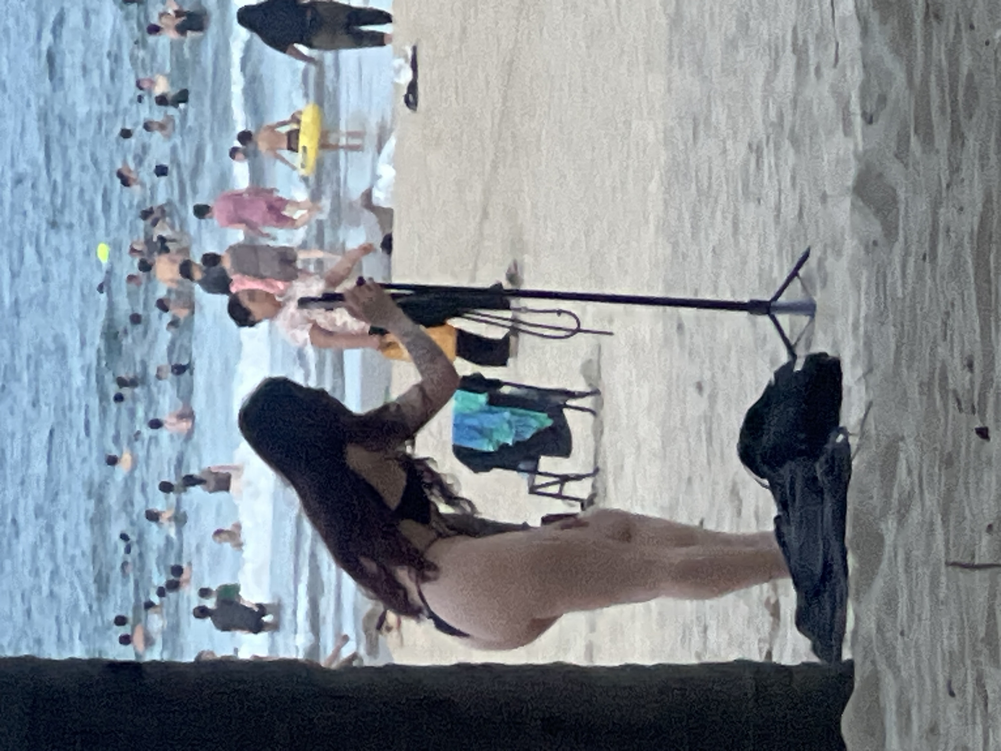 This image depicts a crowded beach scene in Da Nang, Vietnam. In the foreground, a woman in a bikini stands near a dark pole, with a black bag and a tripod on the sand. She has long, dark hair and tattoos on her arms. In the background, numerous people are in the ocean, swimming and wading. The water is blue and appears to be choppy. The beach is sandy, with a few scattered objects. A chair with towels is visible on the sand behind the woman. The overall impression is a busy, recreational day at the beach.