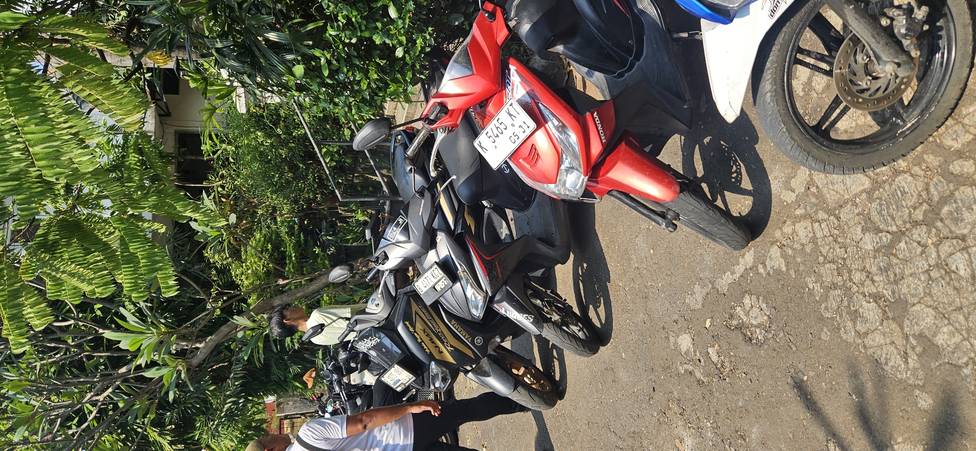 In an outdoor, sunlit scene in Bekasi, Indonesia, a row of parked motorcycles is visible on a paved surface. The motorcycles are predominantly scooters, with a mix of black and red models. One black Yamaha NMAX scooter is prominent in the foreground, displaying a license plate that reads "B 4171 KGZ." Another red Honda scooter is also visible, with a license plate reading "K 5465 KT." Two individuals are present. On the left, a person wearing a white t-shirt and dark pants stands facing away from the camera, their back to the viewer. To the right of the motorcycles, a young person with short dark hair, wearing a light green t-shirt, is looking to the right, away from the parked vehicles. The background features lush green foliage and trees, suggesting a location with ample vegetation. The paved ground appears somewhat worn and cracked.