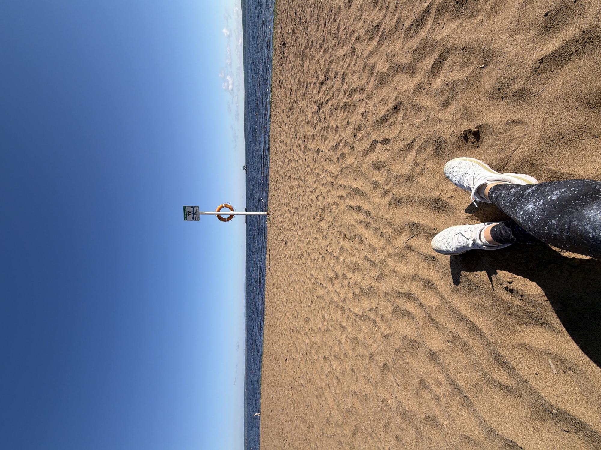 The image depicts a sandy beach with a person's legs and feet, clad in white sneakers and patterned leggings, positioned in the foreground. The sand is textured with footprints and ripples. Beyond the beach lies a body of water, likely a river or lake, with a calm surface reflecting the clear blue sky. In the mid-ground, a white pole stands adorned with a sign featuring two figures and the text "10м", indicating a distance marker. A life preserver ring is attached to the pole. Further out on the water, a ship is visible. The scene appears to be the Kama River embankment in Chaikovsky, Russia.