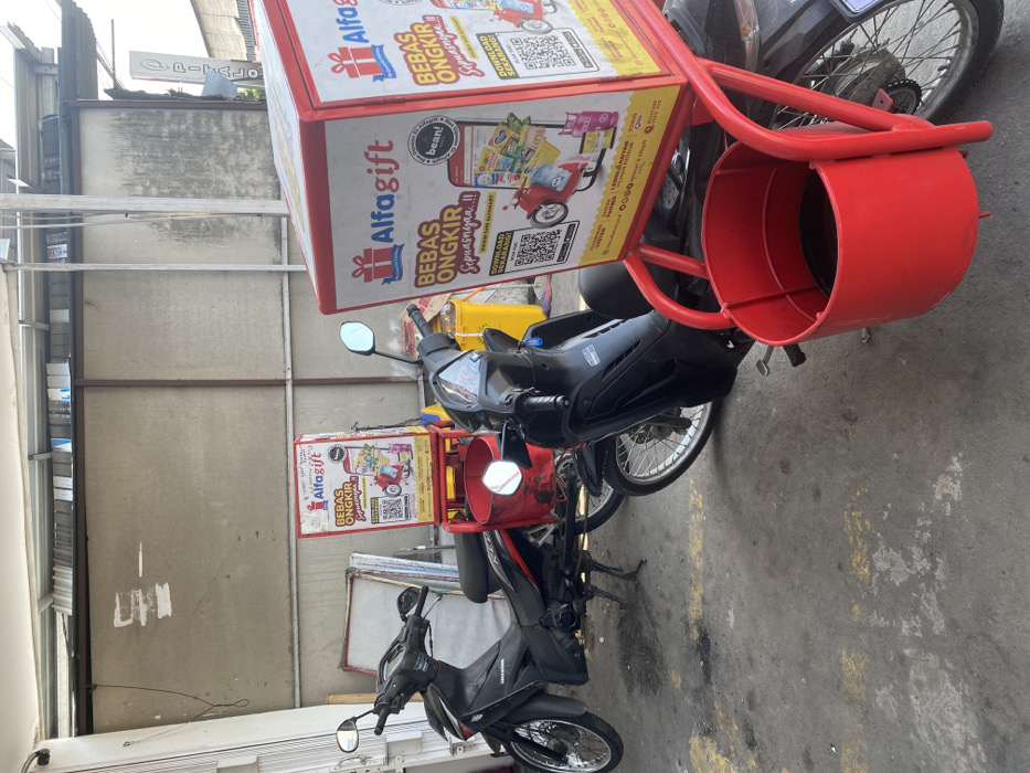 The scene is located in Kecamatan Piyungan, Indonesia, at coordinates -7.824 latitude and 110.48 longitude. The image shows a street corner with two motorcycles parked. Both motorcycles are black and are in good condition. The motorcycle on the left is a Honda. The motorcycle on the right is attached to a red delivery cart. The delivery cart is emblazoned with advertisements for "Alfagift" and promotes "Bebas Ongkir." The cart has a red, cylindrical container, possibly for storing delivery items, located below the advertisements. Behind the motorcycles, a faded brick wall is visible, suggesting a possible alleyway or back entrance. The background appears to be a residential area with a building that seems to be a storefront, possibly a shop or restaurant.
