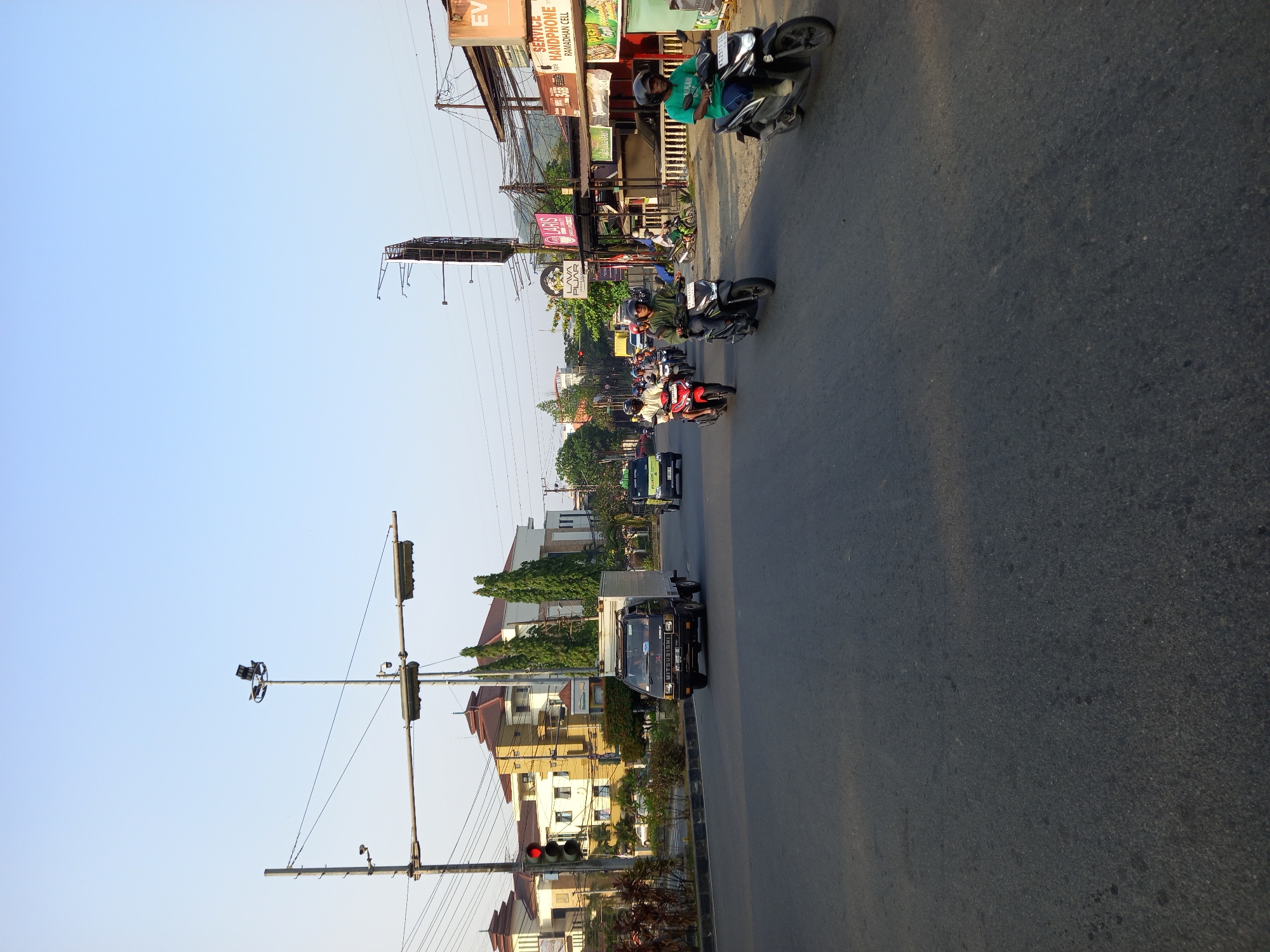 The image depicts a street scene in Samarinda, Indonesia, likely during daylight hours given the clear sky and shadows. The foreground shows an asphalt road with tire marks and scattered debris. Several motorcycles, some with riders, are in motion on the road. In the middle ground, a black Mitsubishi truck is visible, also in transit. Along the sides of the road, buildings, trees, and street infrastructure are present. This includes traffic lights, power lines, street lamps, and various shopfronts with signage. One building appears to be a multi-story structure with cream-colored walls and red-tiled roofs, while another is a more commercial building with awnings and advertisements, one of which reads "RAMADHAN CELL." The overall impression is of a moderately busy urban environment.