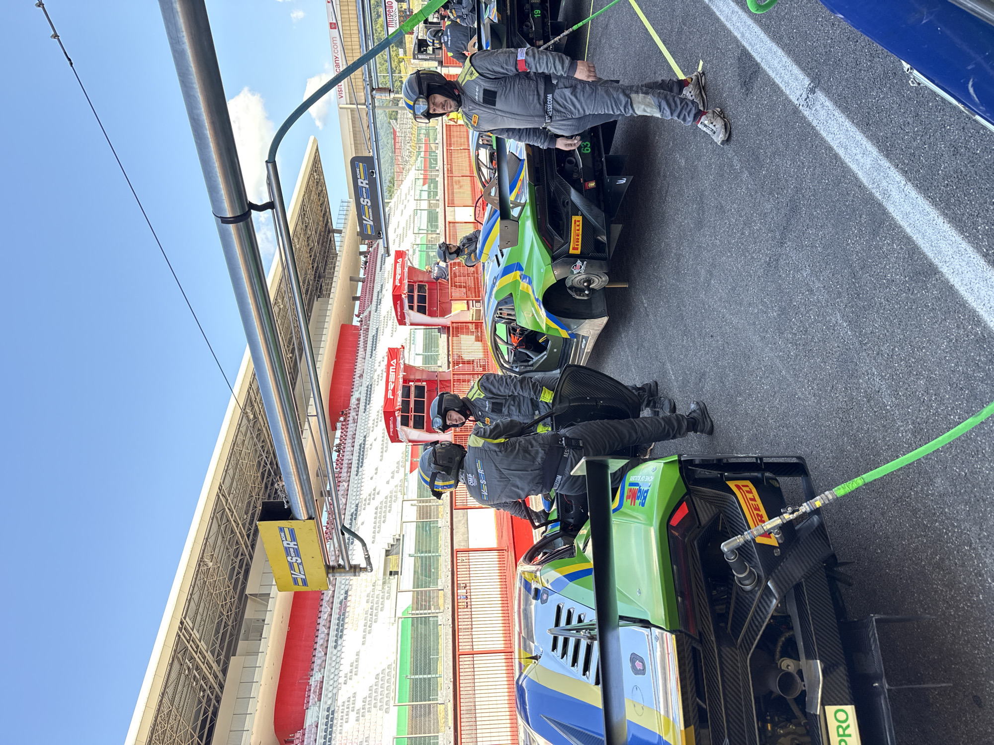 At the Autodromo Internazionale del Mugello, Scarperia, Italy, on a bright, sunny day, a pit stop is underway. Two race cars, a green and blue Lamborghini with the number 63 visible on its rear, and another partially obscured black Lamborghini, are positioned in the pit lane. Several individuals in racing suits and helmets are standing near the cars. One person in a grey suit with "OMP" and "V-S-R" logos is facing the camera, while another in a similar suit stands behind the green car. A green hose extends from the rear of the green car towards the foreground. In the background, the grandstands of the racetrack are visible, with spectators seated. The scene is set against a clear blue sky.