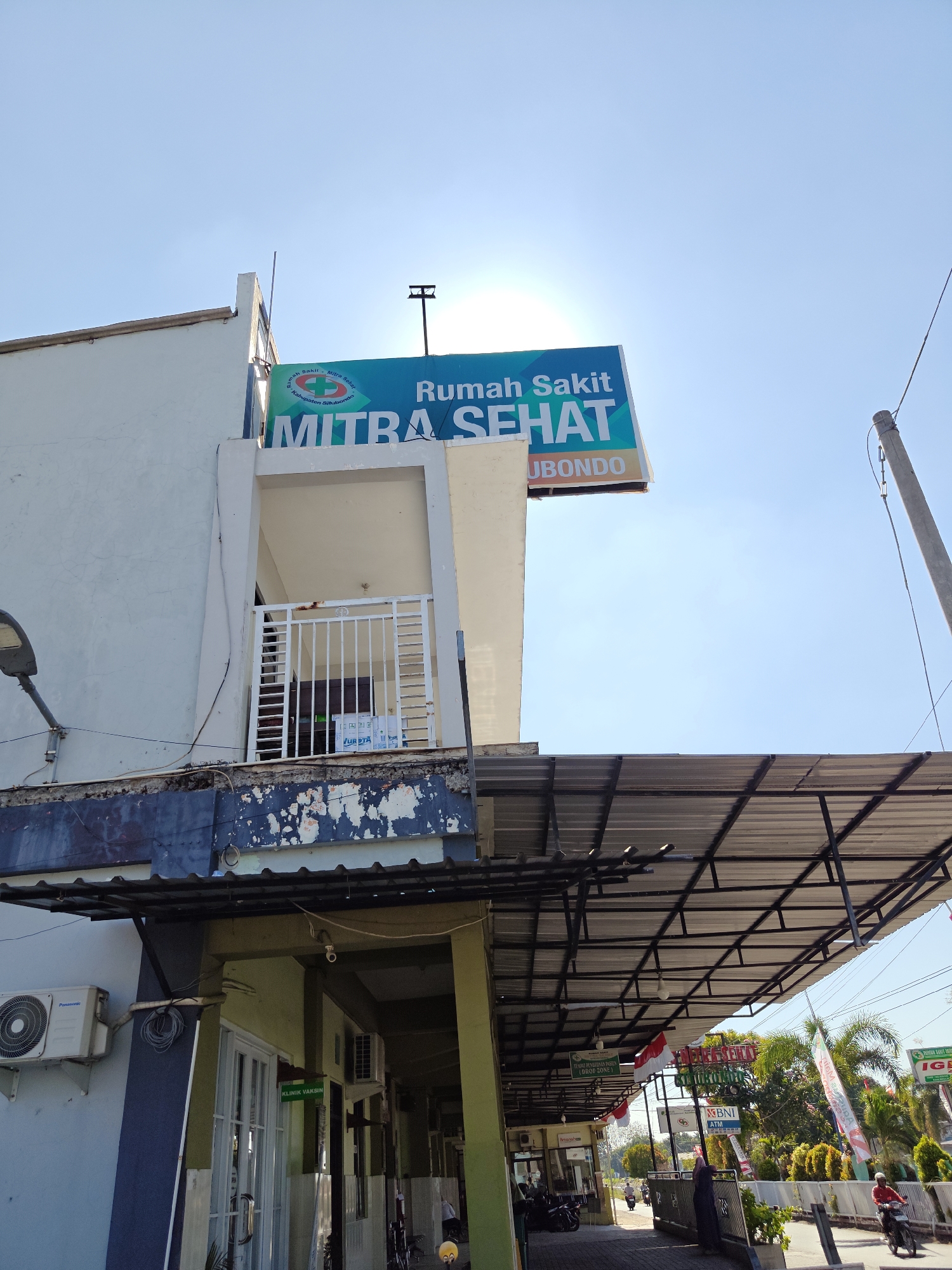 This image shows the exterior of Rumah Sakit Mitra Sehat, a hospital located in Situbondo, Indonesia. The sky is clear and blue, with the sun prominently visible, casting a glare. The hospital building is white with a large teal and white sign that reads "RUMAH SAKIT MITRA SEHAT" and below it, "KABUPATEN SITUBONDO". A covered walkway extends from the building, supported by a metal frame.
On the building itself, there is a balcony with a white metal railing. An air conditioning unit is mounted on the exterior wall. Below the covered walkway, the entrance to the hospital is visible, with doors and windows. A sign reading "KLINIK VAKSIN" (Vaccine Clinic) is attached to the wall near the entrance.
Various signs and banners are visible along the street in front of the hospital, including a BNI ATM sign and a banner with Indonesian flags. Several motorcycles are parked near the entrance, and a few people are present. One person is walking under the covered walkway, and another person is riding a motorcycle on the street. Lush green trees and palm trees are visible in the background, suggesting a tropical environment. The overall scene depicts a typical day at a public hospital.