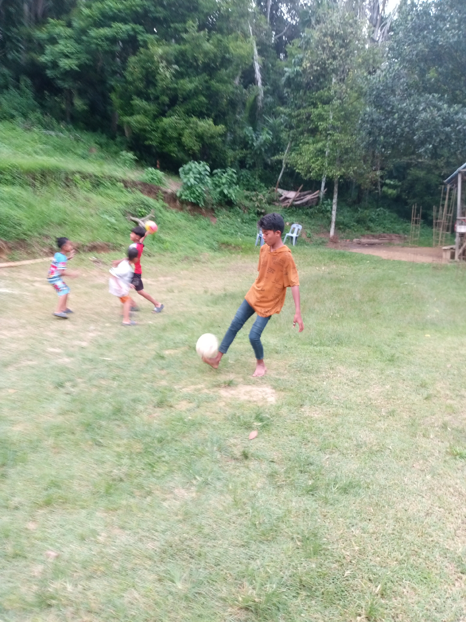 The media file shows a group of children playing soccer in a grassy outdoor area. In the foreground, a young boy in an orange t-shirt and blue jeans is kicking a white soccer ball. To the left, three other children are also engaged in the game, one in a striped shirt and shorts, another in a red shirt and black shorts, and a third in a white shirt and orange shorts. The background features lush green trees and vegetation, suggesting a natural, perhaps rural, setting. A small, rustic structure, possibly a shelter, is visible on the far right. The overall impression is one of casual, outdoor recreation. The lighting suggests it is daytime, possibly late afternoon given the soft light. The environment appears to be a field or a park, typical of a community gathering or play area. There is no visible text in the image.