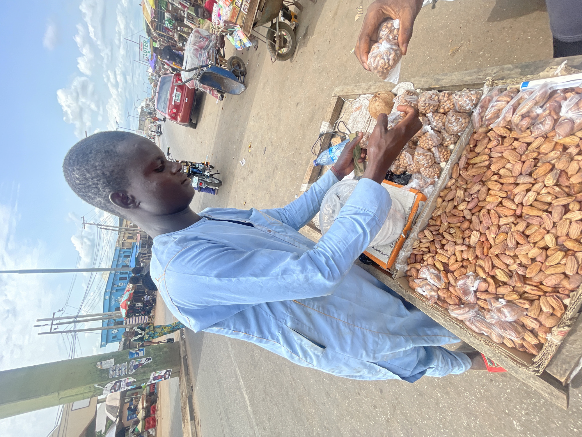 A street vendor wearing a light blue long-sleeved shirt is selling peanuts and dates from a wooden cart in Lokoja, Nigeria. The cart is divided into sections, with dates in one and bags of peanuts in another. Another person's hand is visible holding a bag of peanuts. The street scene in the background includes motorbikes, cars, buildings, and other street vendors. The sky is partly cloudy.