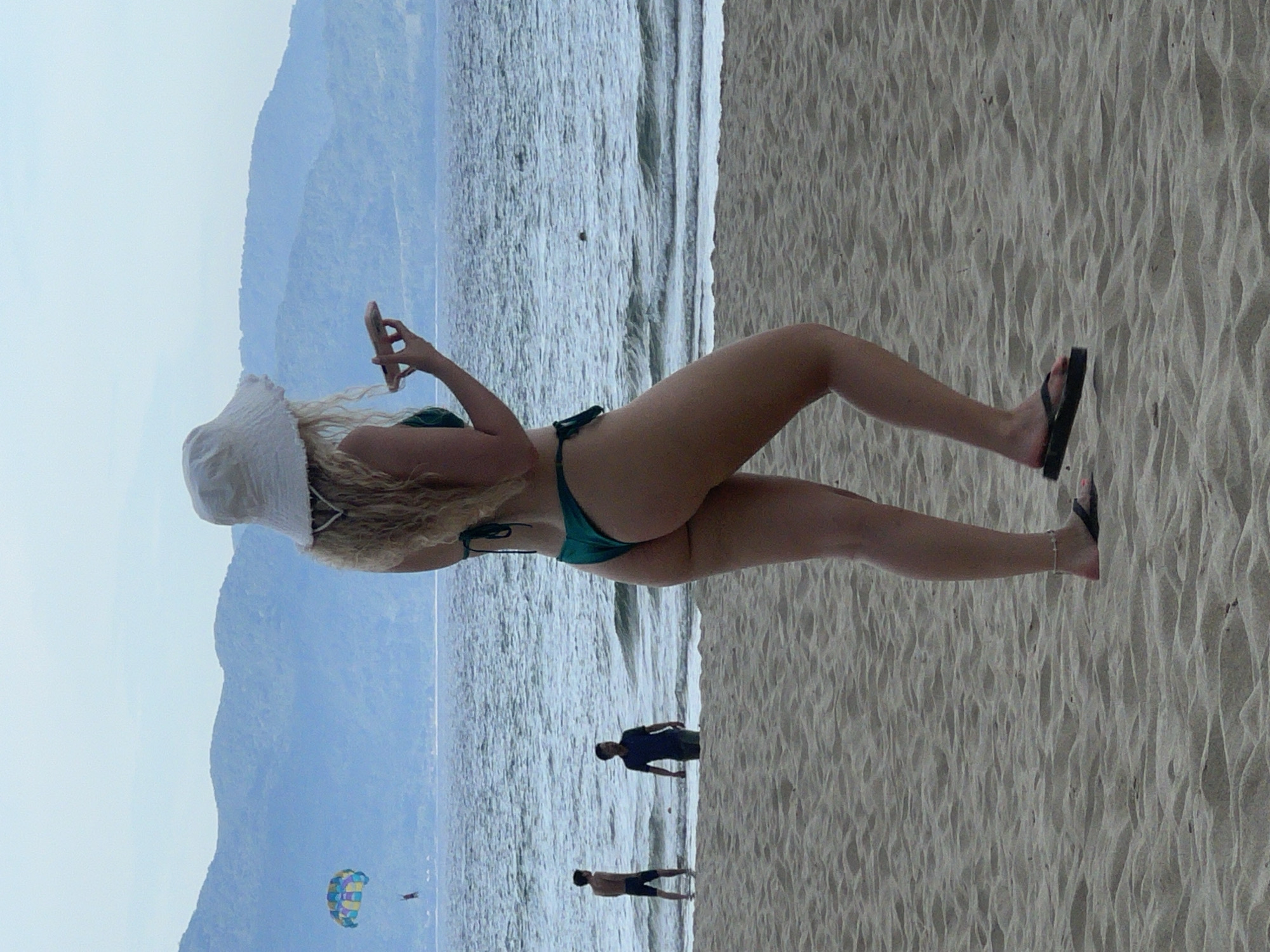 The image shows a young woman in a green bikini and a white bucket hat standing on a sandy beach in Da Nang, Vietnam. She is holding a pink object in her hand and looking out at the ocean. In the background, two men are standing in the shallow water, and a parasailer is visible in the sky. The sea is calm, with small waves rolling onto the shore. Mountains can be seen in the distance, shrouded in a light mist. The sand is textured with ripples, indicating a gentle breeze.