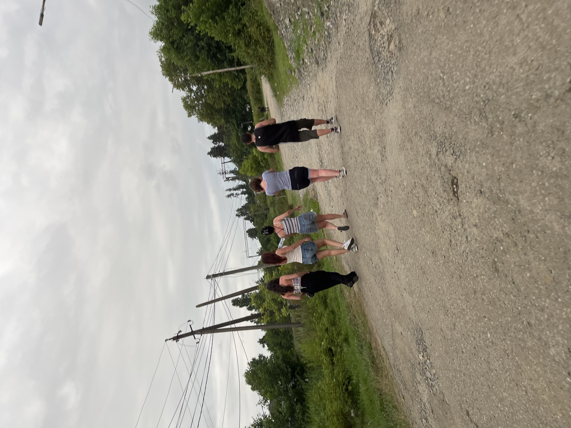 A group of four individuals, three females and one male, are walking away from the camera down a gravel road in Ureki, Georgia. The females are wearing casual attire: two in shorts and tank tops, and one in black pants and a patterned top. The male is wearing a black tank top and olive-colored shorts. The road is surrounded by lush green vegetation and trees, with power lines visible in the background, suggesting a rural or semi-rural setting on the outskirts of the city. The sky is overcast, creating a diffused light.