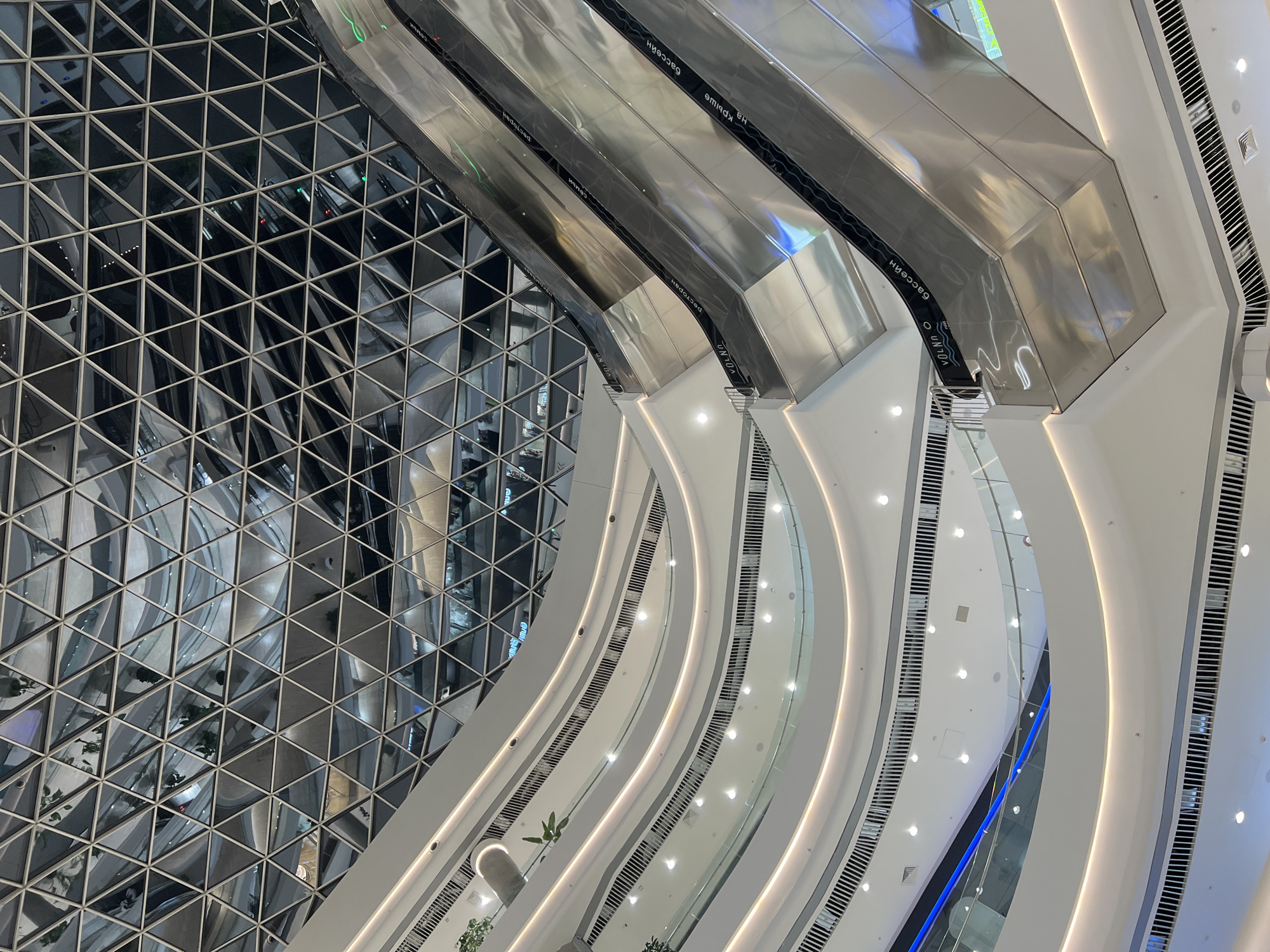 The image captures the interior of a grand, multi-story modern building, likely a shopping mall or a large public atrium, viewed from a high vantage point looking downwards. The scene is characterized by striking contemporary architecture, combining angular and curvilinear forms.
A prominent feature on the left is a vast, reflective wall or ceiling composed of numerous triangular panels, creating a complex geometric pattern. These panels reflect the interior, showing distorted, abstract views of the levels below and ambient lights. Adjacent to this, several sets of escalators ascend diagonally through the space. These escalators are constructed from polished, reflective metal surfaces and feature black handrails.
The right side and bottom of the image reveal multiple curving levels or balconies, stacked vertically. These levels are smooth, white, and brightly illuminated by continuous strips of warm and cool-toned LED lighting embedded along their edges. The design emphasizes clean lines, sleek surfaces, and a sense of openness and upward movement. Small green plants are sparsely placed on some levels, adding a touch of nature to the otherwise minimalist design.
No people are visible, suggesting either an early hour or a less busy moment. The overall ambiance is bright, modern, and spacious, with a predominant color palette of white, silver, and grey, accented by subtle reflections of green and blue lights.
Visible text can be seen on the black handrails of the escalators, often reflected or inverted due to the shiny surfaces. Recognizable Cyrillic words include "БАССЕЙН" (BASSEYN, meaning "swimming pool"), "РЕСТОРАН" (RESTORAN, meaning "restaurant"), and "НА КРЫШЕ" (NA KRYSHYE, meaning "on the roof"), indicating services or destinations within the building. English text like "VOL NO." also appears.
The setting is a large interior space in Khabarovsk, Russia, showcasing a blend of functionality and sophisticated design.