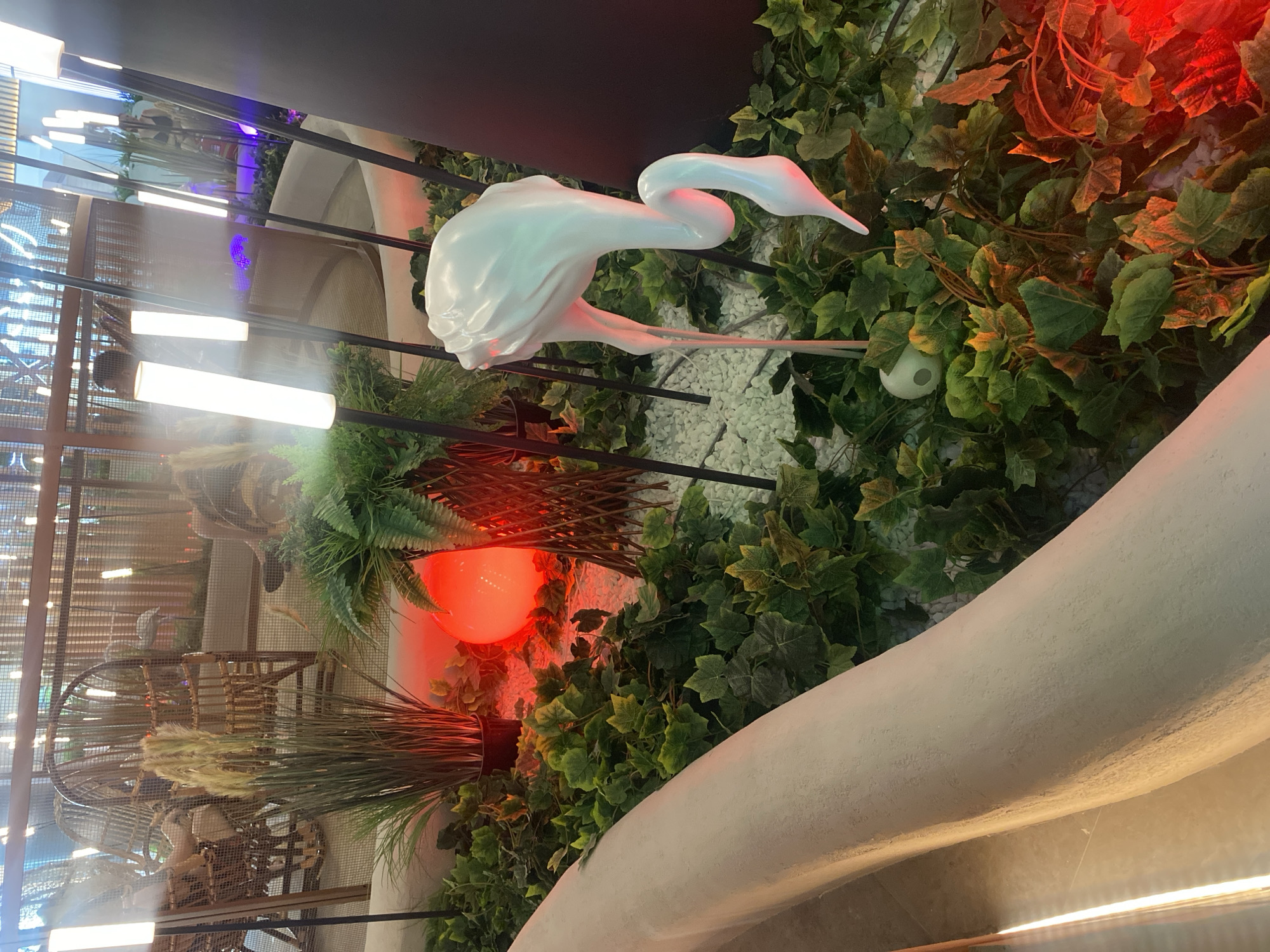 The image depicts an indoor decorative scene featuring lush green artificial foliage, white gravel, and several illuminated cylindrical lamps. A large, white, abstract sculpture resembling a flamingo with a long, curved neck and legs is the central subject. The flamingo sculpture is positioned amongst the plants. In the background, wicker chairs are visible, suggesting an indoor seating area or lounge. A large, glowing red orb is also present, casting a warm light on the surrounding greenery. The lighting in the scene is a mix of cool white from the lamps and the warm red from the orb, creating a dramatic atmosphere. The overall impression is of a modern, artfully designed interior space. The image does not contain any elements that would allow for the specific location in Berdsk, Russia, to be determined.