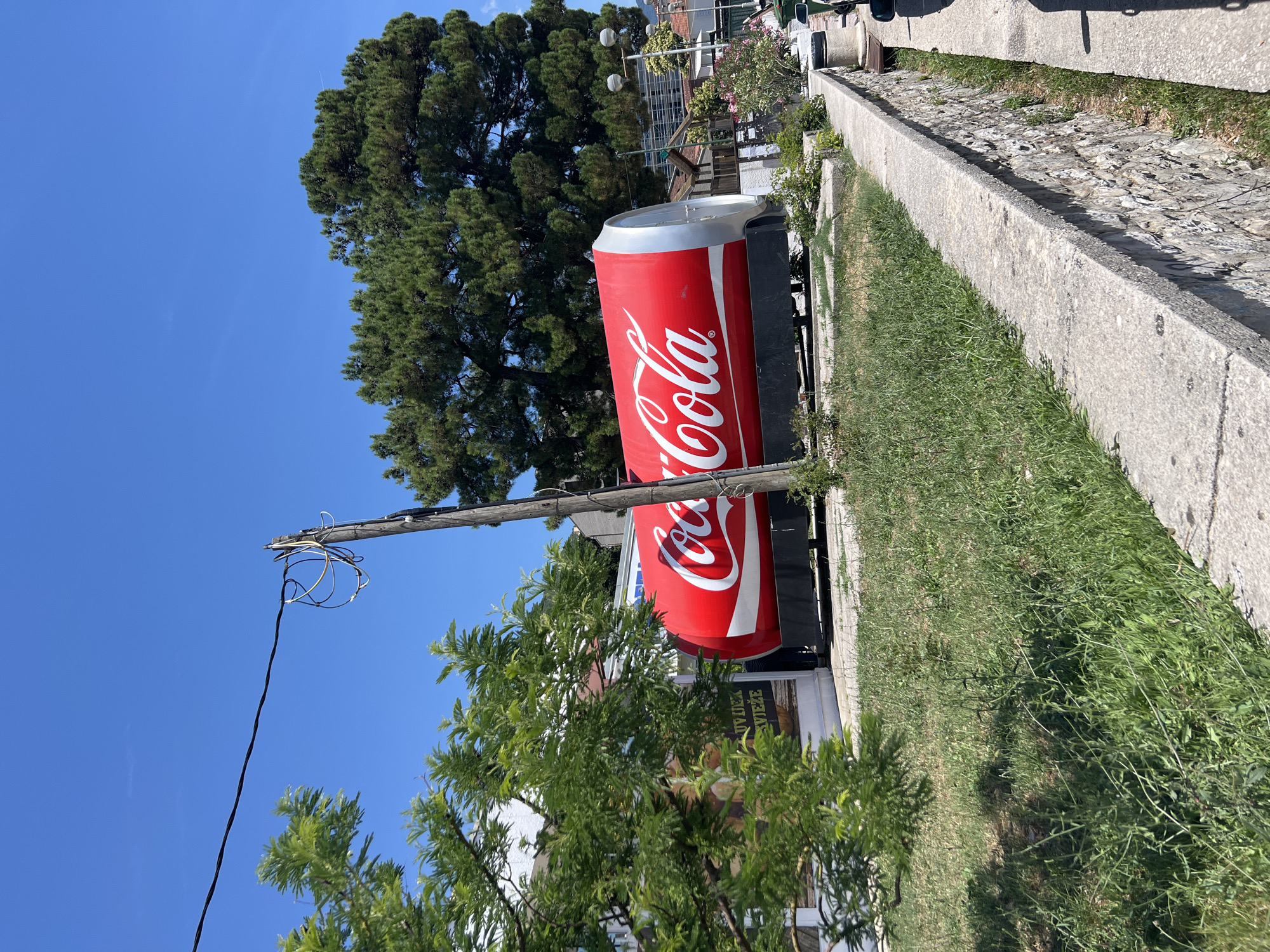 The image shows a large, red Coca-Cola can shaped structure situated outdoors in what appears to be a public space in Baošići, Montenegro. The structure is the dominant subject, with the Coca-Cola logo clearly visible in white script. It is lying horizontally, supported by a dark base. To the left, there are green trees and a building with some signage. A wooden utility pole with wires is prominent in the foreground and middle ground, to the left of the Coca-Cola can. Behind the can, a large, mature green tree provides a natural backdrop. The ground is covered with grass and some low-lying vegetation, and a concrete wall with stone construction underneath runs along the right side of the image, suggesting a pathway or elevated area. The sky is clear and blue.