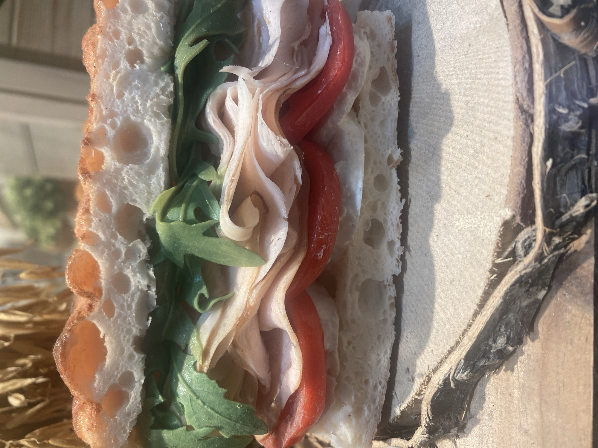 A close-up, top-down shot depicts a sandwich, likely a bocadillo, with visible layers of toasted bread, green arugula, thinly sliced pale meat (possibly turkey or chicken), and red peppers. The sandwich rests on a cardboard or wooden surface, with fragments of straw or wood shavings visible to the right. The background is slightly blurred, hinting at an outdoor location, possibly a café or picnic setting. The light suggests it is daytime. Specific location in Barcelona is not determinable from the image.