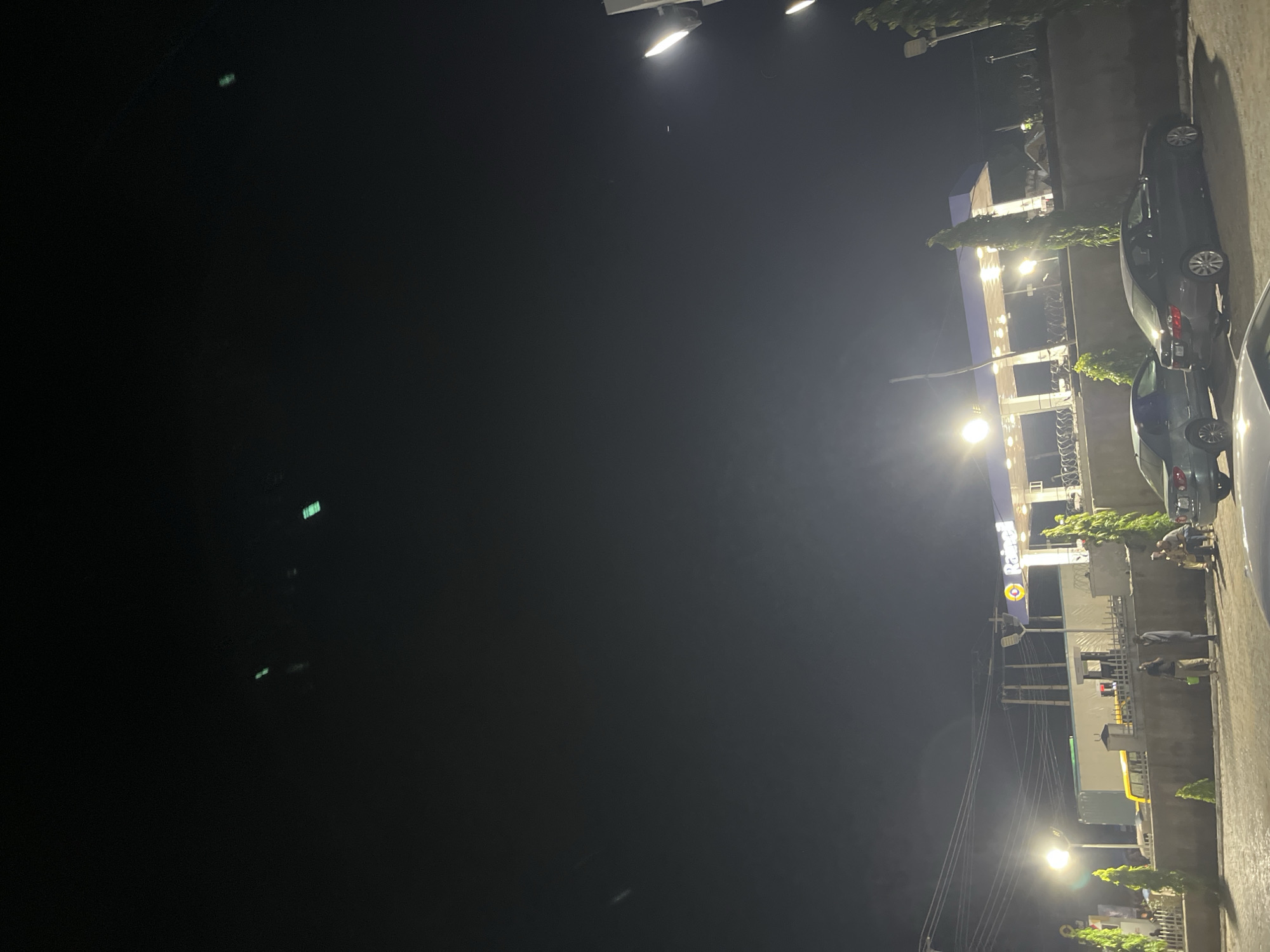 The image depicts a nighttime scene at a gas station in Jalingo, Nigeria. A "Rainoil" branded canopy is illuminated by bright lights, with a series of fuel pumps visible beneath it. Several cars are parked in front of the station. In the foreground, a low concrete wall separates the parking area from the street. Some people are visible standing and sitting near the wall. One person is holding a green bucket. A yellow truck is parked behind the wall. The sky is dark, with some distant lights and a few faint, bright streaks that could be aircraft or atmospheric phenomena. The overall impression is a typical roadside scene at a gas station after dark.