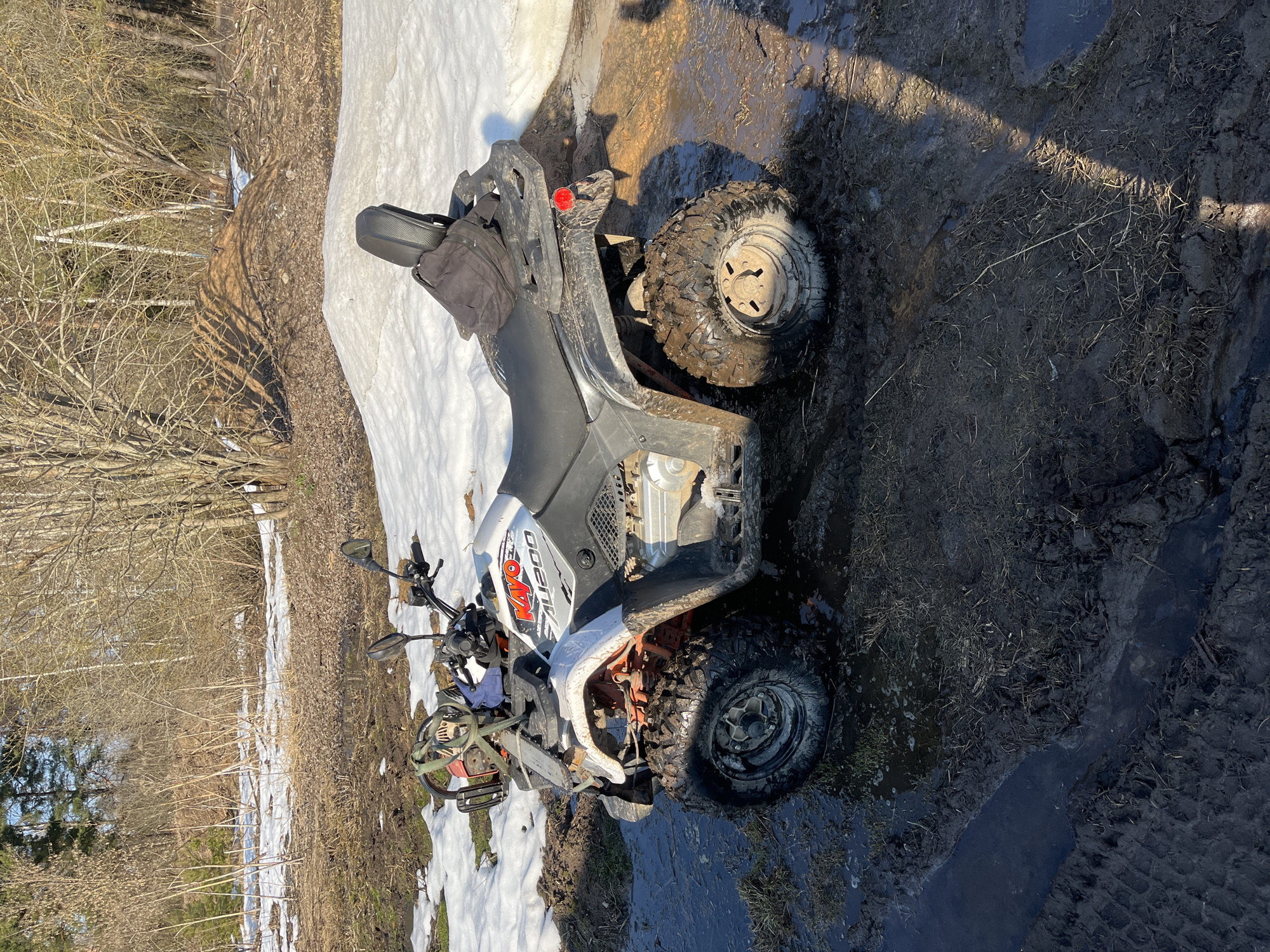An outdoor scene in Razdol'e, Russia, depicts a stationary Kayo AU200 all-terrain vehicle (ATV) on a muddy, unpaved surface. The white and black ATV is heavily caked with dark brown mud on its tires, wheels, suspension, and lower body components. A red and silver chainsaw is secured horizontally to the front cargo rack. A black bag rests on the rear cargo rack, positioned against a black backrest. The ground consists of saturated dark brown mud, standing water puddles, and patches of melting white snow, exhibiting distinct tire tracks. Sparse dry grass and brown foliage are interspersed within the terrain. The background reveals a forest edge characterized by bare deciduous trees and shrubs under clear skies, with some evergreen trees visible further in the distance. A small mound of disturbed earth or debris is present in the mid-ground. The scene is illuminated by direct sunlight, casting shadows. No individuals are present.