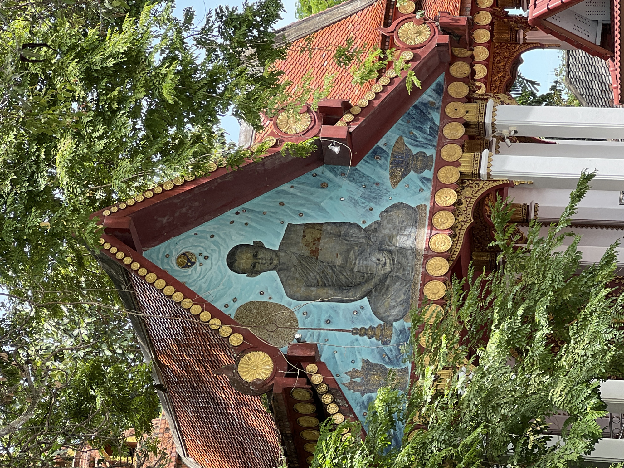 This media file captures a close-up, angled view of the exterior of a building, likely a temple or shrine, in Baan Hua Thanon, Thailand. The building features traditional Thai architecture with a steeply pitched, orange-tiled roof edged with decorative gold circular motifs. The focal point is a mural painted on an angled section of the building's facade.
The mural depicts a serene, meditating figure, likely a monk, rendered in muted grey tones against a bright, turquoise blue background. The figure is seated, with hands clasped, and appears to be floating or suspended. Surrounding the figure are abstract patterns and shapes, including what appear to be stylized fish and a circular emblem with radiating lines. A string of gold circular ornaments runs along the roofline above the mural.
Lush green foliage from nearby trees encroaches on the scene, partially obscuring the building and its decorative elements. The lighting suggests it is daytime, with sunlight filtering through the leaves. The overall impression is one of cultural artistry and spiritual tranquility, set within a tropical environment. No visible text is present in the image.
