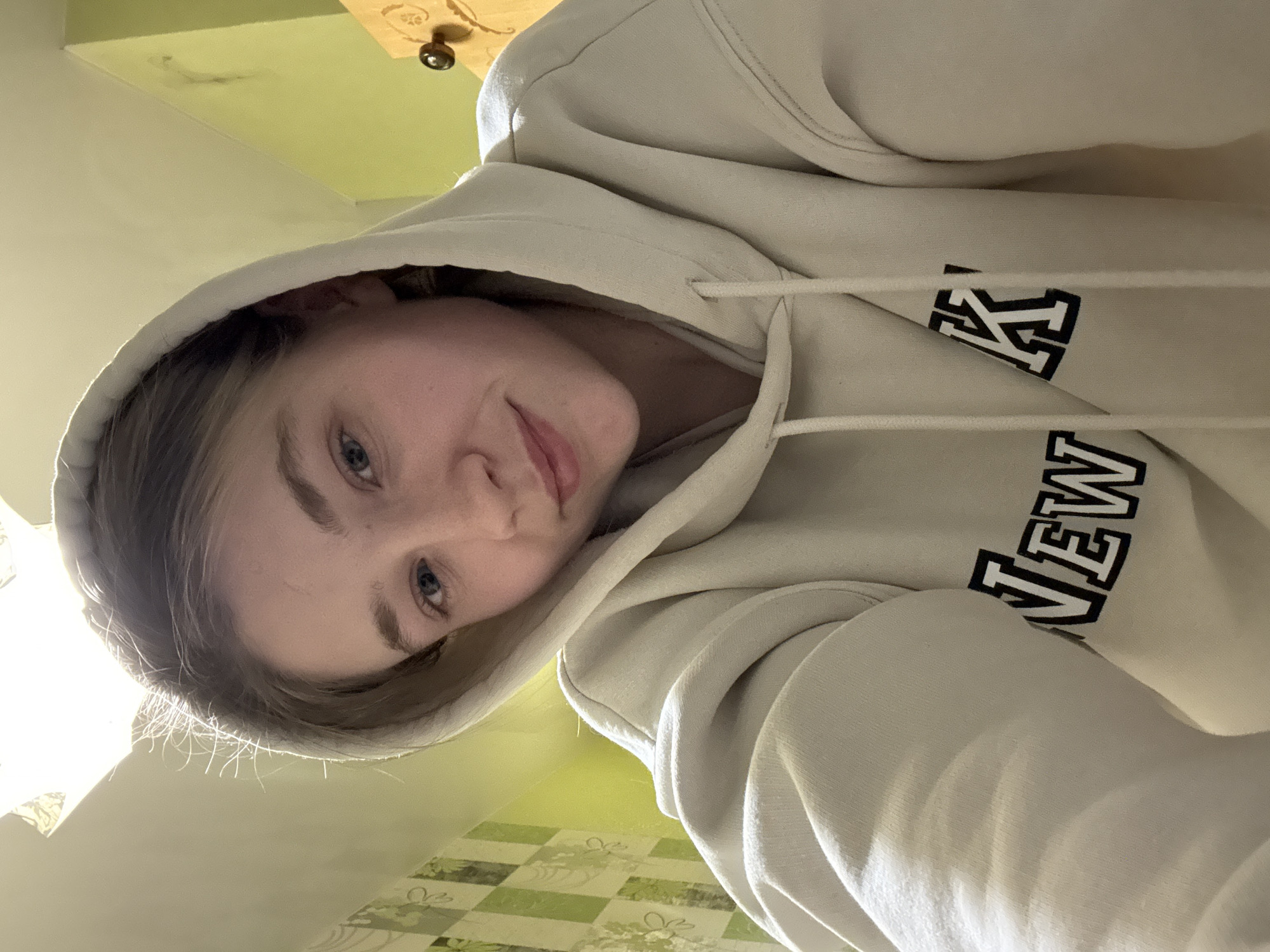 A young woman with fair skin and blue eyes is wearing a beige "NEW YORK" hoodie. She is looking down at the camera with a slight smile. The background includes a light green wall with a patterned surface and a piece of furniture with a drawer pull. The location in the city of Izhevsk, Russia is not discernable from the visual information.