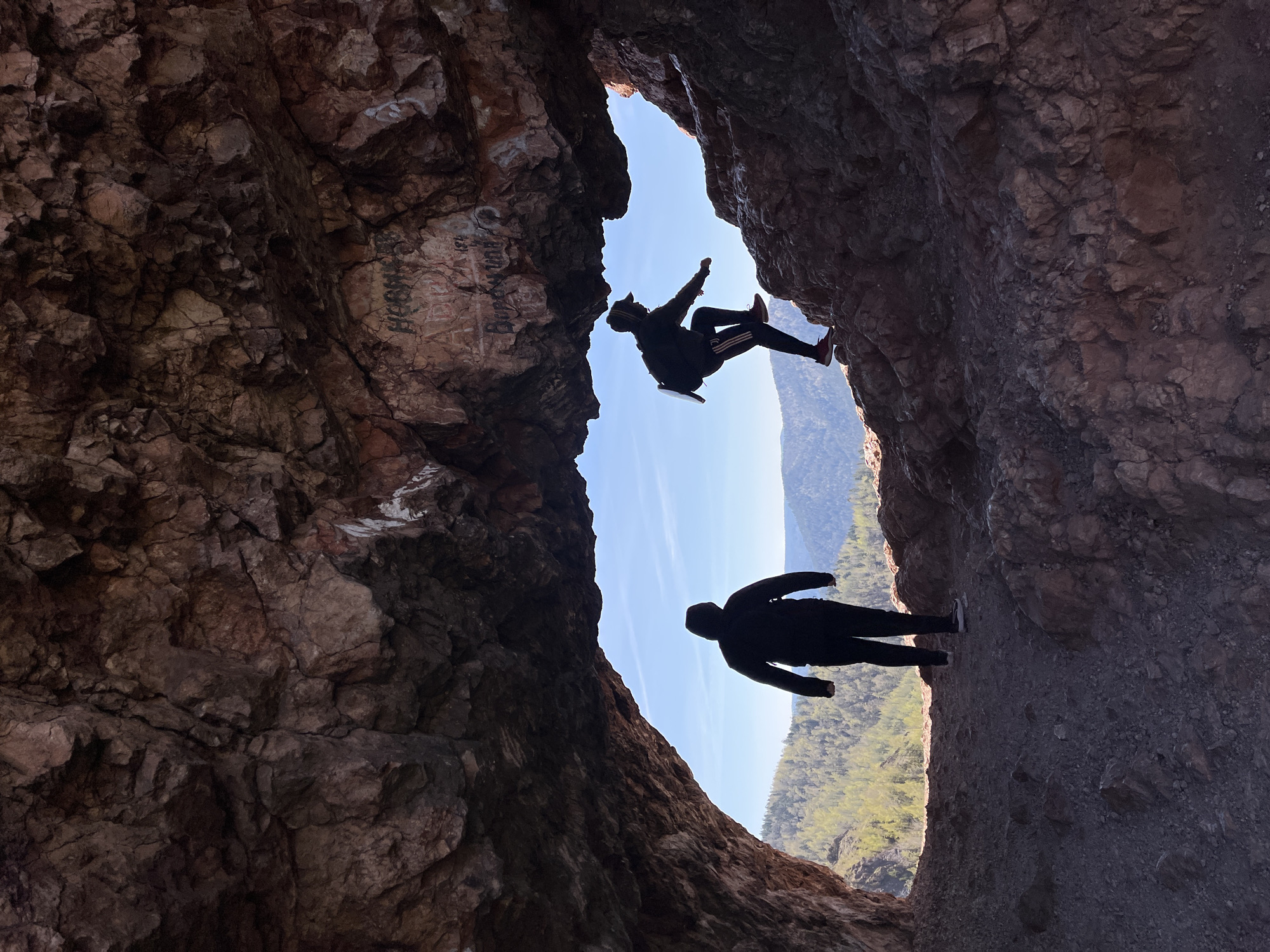The image shows two people silhouetted against a bright sky, framed by the rock formations of the Tsar-Vrata rock in Krasnoyarsk, Russia. One person is positioned high up in the rock opening, seemingly attempting to climb or traverse across, with one arm extended and legs bent. A backpack is visible. The other person is positioned lower down in the opening, standing. The surrounding rock is textured and shows signs of weathering, with some graffiti present on the upper portion. The background shows a valley with trees.