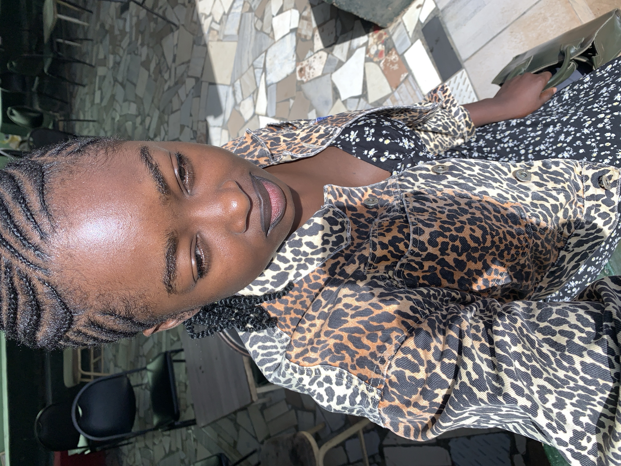 A young Black woman is depicted in what appears to be an outdoor or semi-outdoor setting, likely a patio or courtyard in Kabong, Nigeria. The image is rotated 90 degrees clockwise. She is seen from the chest up, looking slightly upwards with a gentle smile. Her face is well-lit, highlighting her features. She has dark, neatly styled cornrows on the side of her head.
She is wearing a fashionable leopard print jacket over a dark dress with a small floral pattern. Her right hand is visible, holding a green handbag resting on her lap. The background features a distinctive tiled floor composed of irregularly shaped light and dark tiles, creating a mosaic-like pattern. Strong shadows are visible on the ground, indicating bright daylight, suggesting it is midday or early afternoon. In the background, dark metal chairs can be partially seen. No visible text is present in the image.