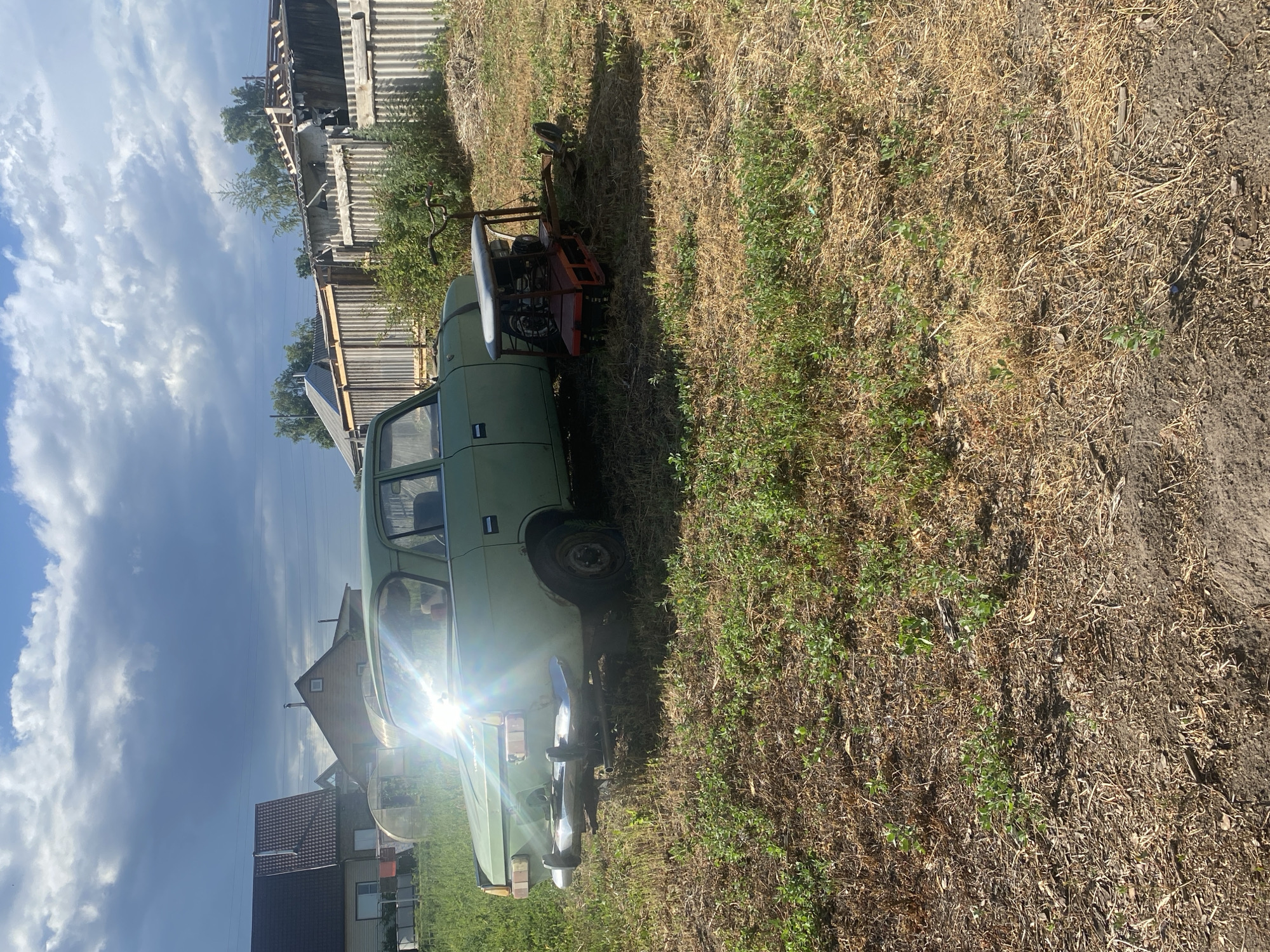 A light green, vintage sedan sits parked in a dry, grassy yard in Mamontovo, Russia. The car is positioned on the left side of the frame, facing right, with its rear towards the foreground. A small, red, mechanized garden tiller with a metal seat and handlebars is attached to the rear bumper of the car, suggesting it may have been towed or modified. The car shows signs of age, with some rust visible on the bumper and body.
In the background, several wooden fences and residential structures are visible. The sky above is partly cloudy, with bright sunlight breaking through, casting lens flare on the car's windshield. The ground is covered in dry grass and some patches of green weeds, indicating a warm, possibly dry, season. The overall scene depicts a rural or semi-rural setting.