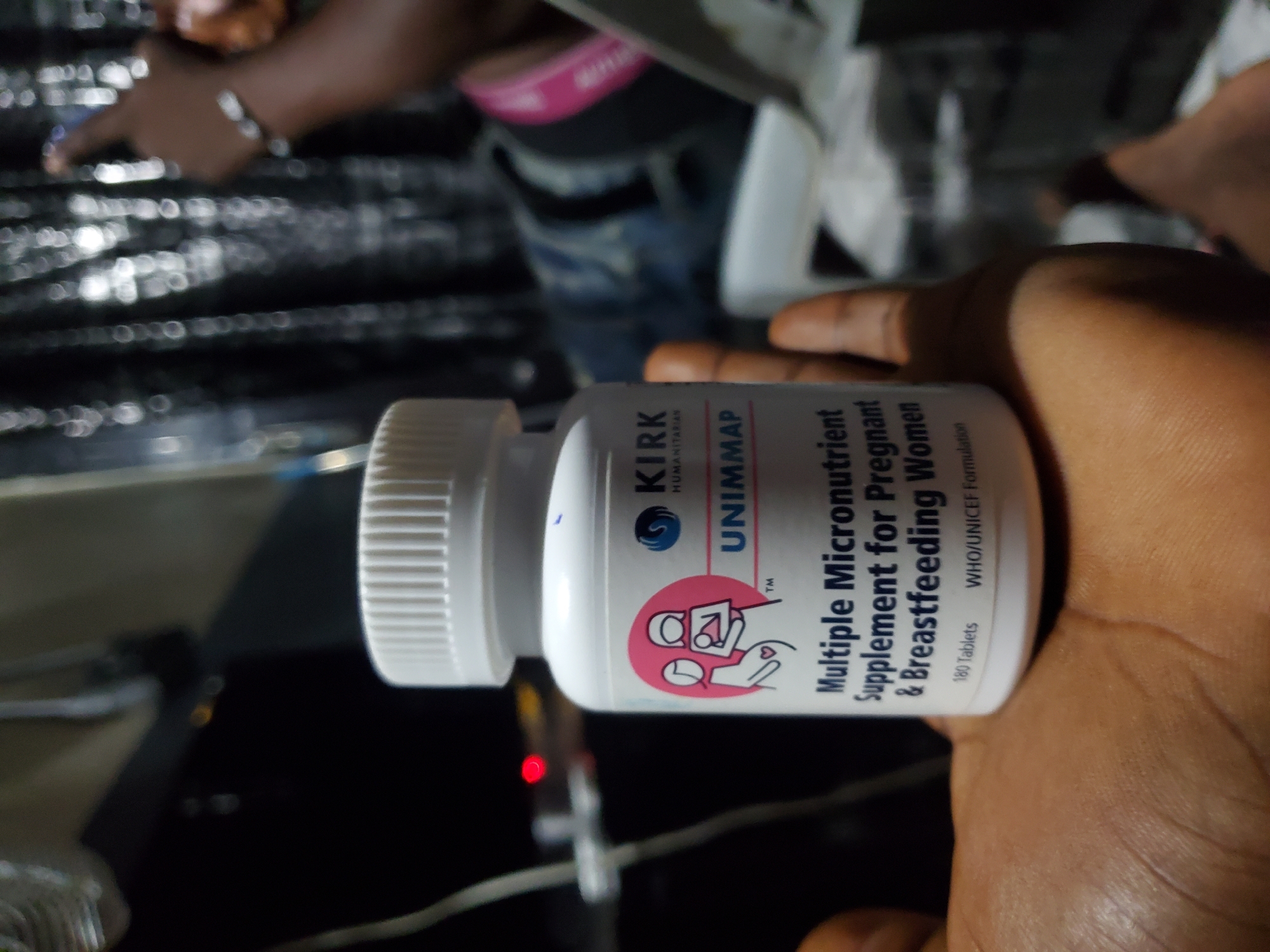This image, captured in the city of Winters, United States of America, depicts a close-up of a brown-skinned hand holding a white plastic bottle. The bottle features a ribbed white screw cap and a label for "KIRK HUMANITARIAN UNIMMAP," identified as a "Multiple Micronutrient Supplement for Pregnant & Breastfeeding Women." The label also states "180 Tablets" and "WHO/UNICEF Formulation," with a pink circular emblem illustrating a pregnant woman and a woman holding an infant.
The background is dark, heavily out-of-focus, and appears to be an indoor setting with low ambient lighting. To the left of the bottle, a thin white cable is visible, alongside a blurred red LED light and the indistinct outline of a dark object. To the right, another brown-skinned arm is partially visible, bent at the elbow, with fingers extended. This arm displays a light-colored wrist bracelet and a pink band on what appears to be clothing. Numerous circular bokeh light effects are scattered across the dark background, suggesting reflective surfaces or distant light sources.