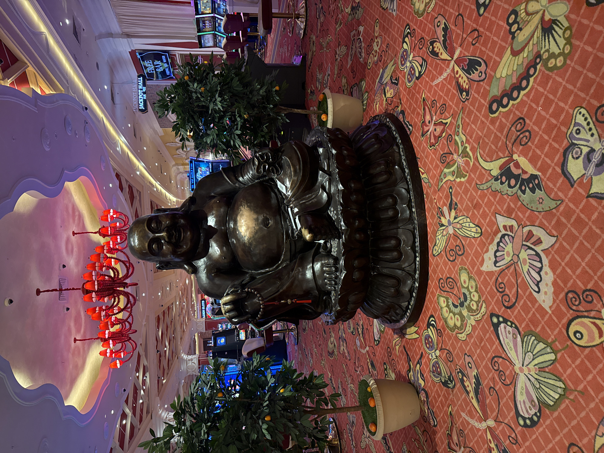 A bronze Buddha statue is located on a red carpet with butterfly designs. The Buddha is surrounded by two small trees in white pots with artificial oranges. In the background, there is a red chandelier hanging from a white ceiling and slot machines are visible. The image appears to be taken indoors in a casino or hotel. The location Shkotovo, Russia, is not visually confirmed in the image.