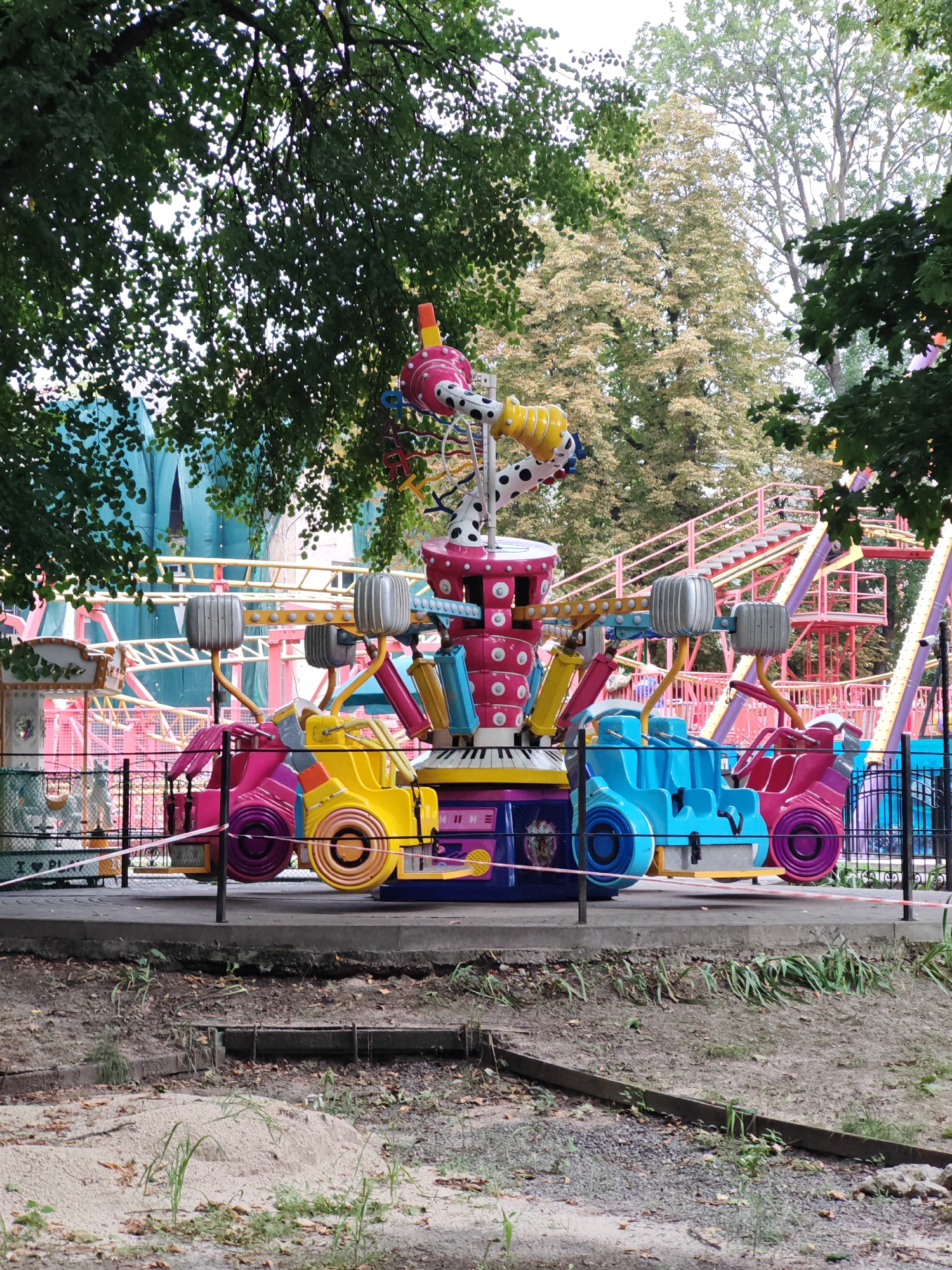 The image depicts a brightly colored amusement park ride, likely a children's ride due to its small scale and whimsical design, situated in an outdoor setting. The ride features multiple seats designed to resemble cars, colored in pink, blue, and yellow. The central column of the ride is adorned with polka dots and various colorful cylindrical elements. In the background, a roller coaster track with pink and yellow supports is visible, suggesting the ride is part of a larger amusement park. Lush green trees with dappled sunlight filtering through their leaves frame the scene. The ground in front of the ride is paved with concrete, and beyond that, a patch of soil with sand and scattered leaves is visible. The scene appears to be a typical amusement park environment, potentially located in Homyel, Belarus, given the context. No people are visible in the image.