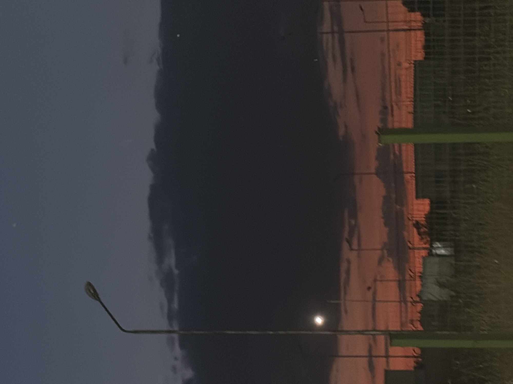 A vertical, wide-angle, low-light outdoor photograph captures a twilight scene. In the foreground, a weathered, green metal fence post stands on the left, with a wire mesh fence extending to the right. Tall, dry grass grows in front of the fence. Behind the fence, a line of industrial-looking buildings or shipping containers is visible, some with reflective surfaces catching faint light. In the mid-ground, several streetlights are illuminated, their poles silhouetted against the sky. One prominent streetlight with an arched arm stands on the left side of the image.
The sky dominates the upper portion of the image. It transitions from a deep, dark blue at the top to a vibrant orange and red hue near the horizon, indicative of sunset or sunrise. Dark, heavy clouds are present in the upper sky, contrasting with lighter, scattered clouds that catch the colorful light closer to the horizon. A few small, bright dots, likely stars or distant lights, are visible in the dark blue expanse of the sky. The overall atmosphere is dusky and somewhat desolate. The location is specified as Aidemir, Bulgaria.