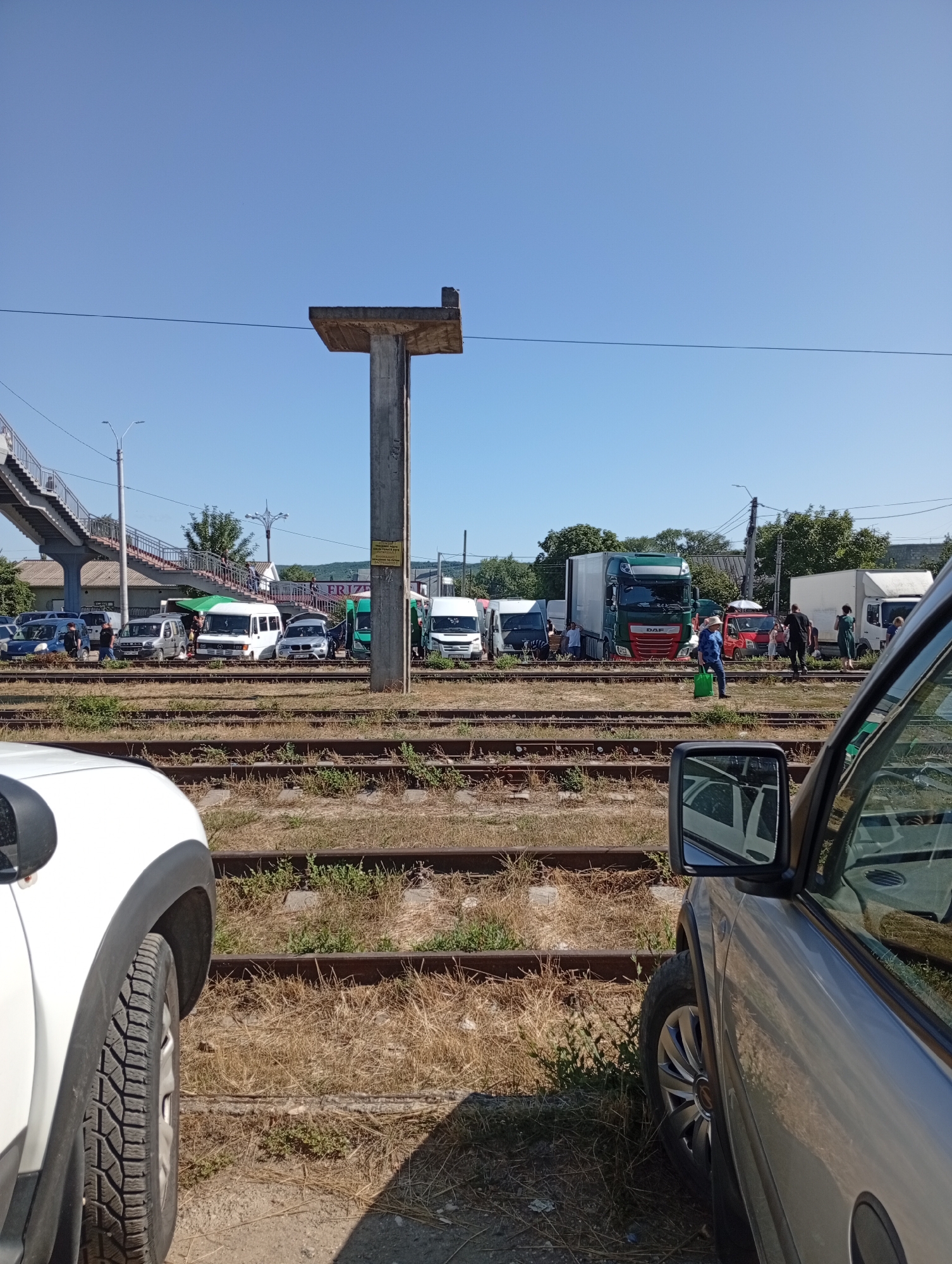 The image captures a scene at a railway station in Strășeni, Moldova, on a clear, sunny day. In the foreground, the side of a white car is visible on the left, and the side of a silver car is visible on the right, with its side mirror reflecting another vehicle. Train tracks run horizontally across the midground, partially obscured by dry grass. Beyond the tracks, a variety of vehicles are parked, including several vans, trucks, and cars. A pedestrian overpass, a concrete structure with stairs, is prominent on the left side of the frame, extending upwards. A tall, unfinished concrete structure, possibly a pylon or support beam, stands near the center of the image, with a crossbar at the top. Several people are scattered among the vehicles and near the tracks. One person in a blue shirt and hat walks with a green bag, while others stand in groups or walk. The background consists of trees and buildings, suggesting a semi-urban or rural setting. The overall impression is one of activity at a transport hub, with vehicles and people present around the railway infrastructure.
