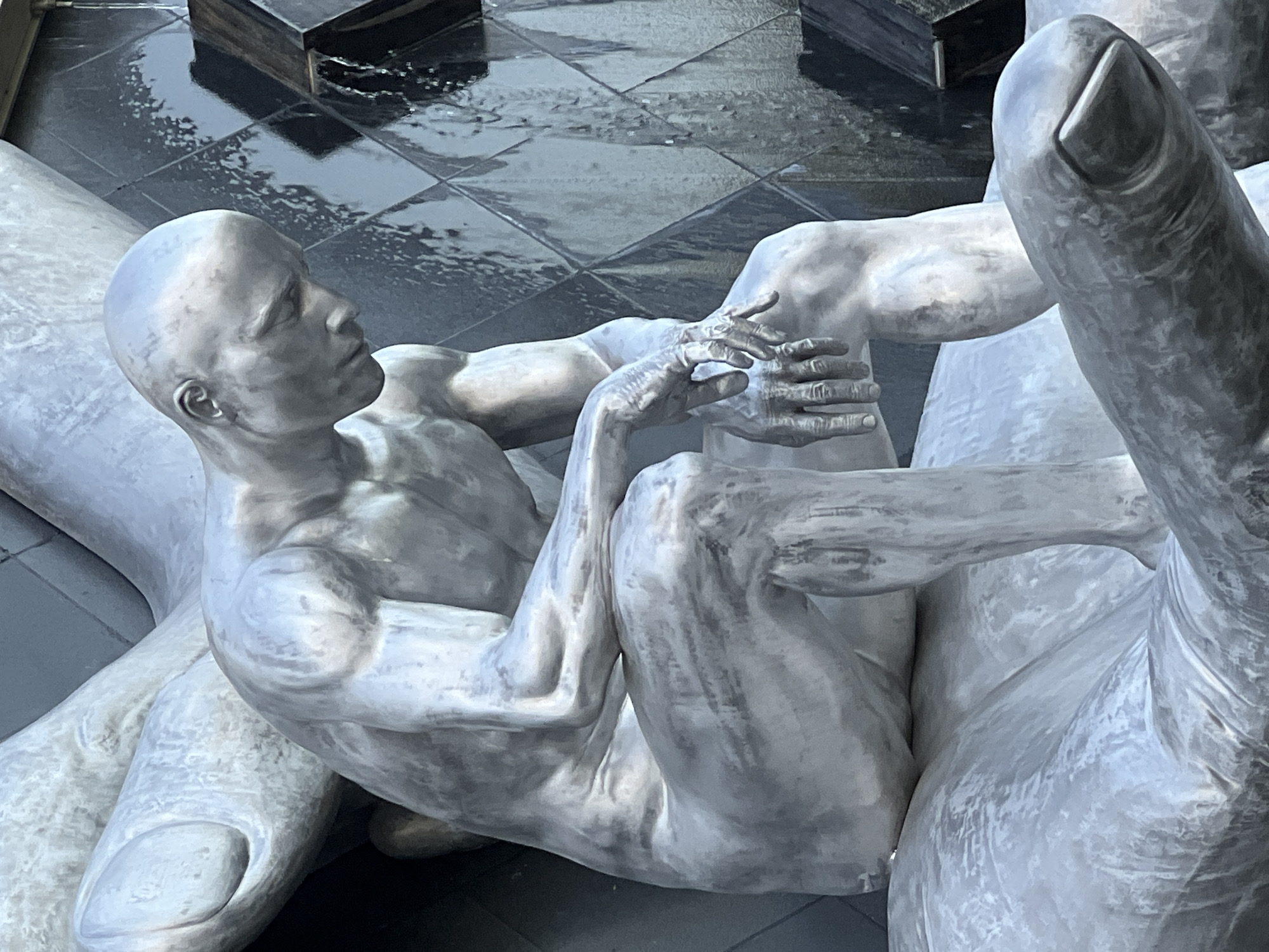 The image shows a close-up of a silver-colored sculpture of a bald man sitting in a pensive pose. He is leaning forward, with his head bowed and his hands resting on his knees. The sculpture appears to be made of metal and is textured to resemble human skin. In the background, a dark, tiled surface is visible with water flowing down it, suggesting it might be part of a fountain or water feature. The sculpture itself is detailed, with visible musculature and veins on the man's arms and legs. The overall impression is one of deep thought or contemplation. The location is the "The Creator" sculpture by Zurab Tsereteli, located in Krasnodar, Russia.