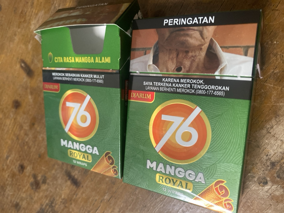The image displays two packs of Mangga Royal 76 kretek cigarettes by Djarum, oriented diagonally on a brown wooden surface. Both packs are predominantly green with a central orange and gold circle containing the number "76" and the text "MANGGA ROYAL". Each pack indicates "12 WRAPS".
The upper pack is partially open at the top, revealing a grey interior flap. Its visible side features a black band with Indonesian text: "DIARUM" and "MEROKOK SEBABKAN KANKER MULUT LAYANAN BERHENTI MEROKOK (0800-177-6565)" (Smoking causes oral cancer. Quit smoking service (0800-177-6565)). Adjacent to this is green text stating "CITA RASA MANGGA ALAMI" (Natural Mango Flavor).
The lower pack is closed and presents a graphic health warning on its side. This warning shows a close-up image of a person with a visible surgical stoma on their throat. Accompanying Indonesian text reads: "PERINGATAN" (WARNING) vertically, and horizontally, "DIARUM", "KARENA MEROKOK, SAYA TERKENA KANKER TENGGOROKAN LAYANAN BERHENTI MEROKOK (0800-177-6565)" (Because of smoking, I have throat cancer. Quit smoking service (0800-177-6565)).
No individuals are present in the scene. The wooden surface exhibits a distinct grain pattern. The scene is located in Cimenyan, Indonesia.