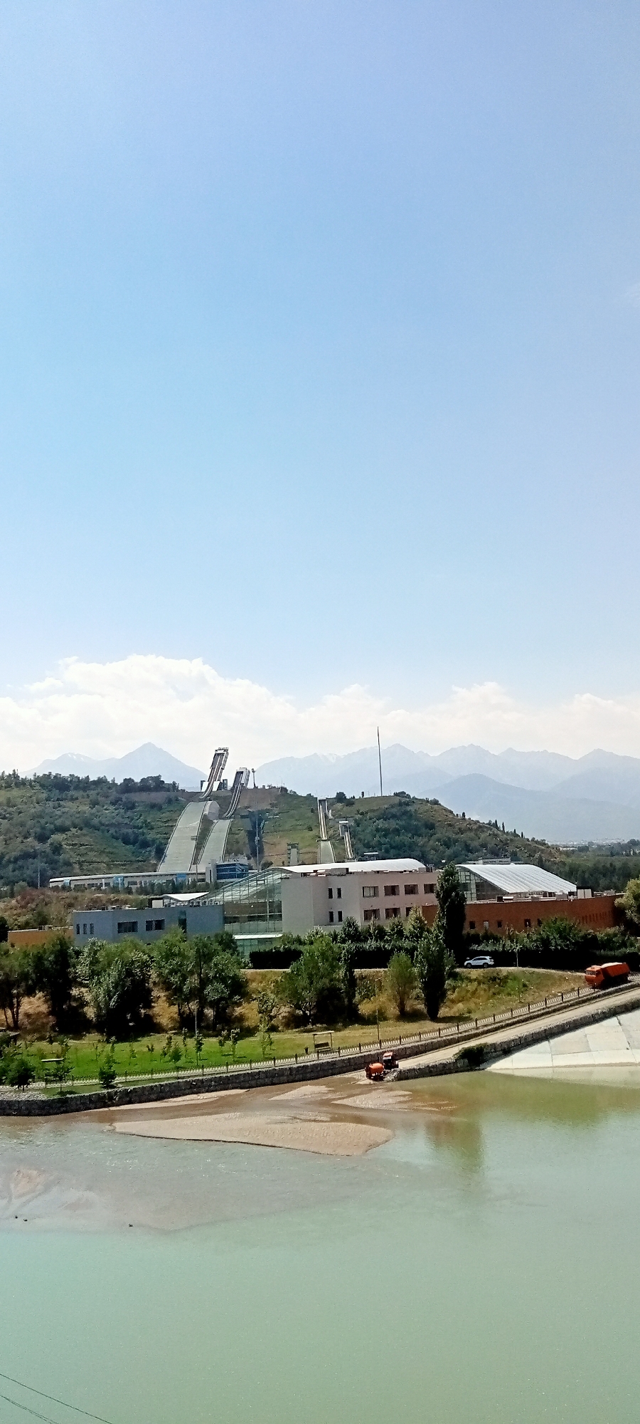 A wide shot captures the Sunkar International Ski Jumping Complex in Almaty, Kazakhstan, on a clear, bright day under a pale blue sky. The complex is nestled against a backdrop of lush green hills and distant, snow-capped mountains. The ski jumps, with their distinctive white runways and supporting structures, dominate the upper portion of the image. Below the jumps are several modern buildings, one with a distinctive glass-covered section, surrounded by green trees and landscaping.
In the foreground, a wide, shallow river or body of water with a pale green hue flows across the bottom of the frame. The riverbed is partly exposed, revealing sandy areas. A paved pathway runs alongside the river, bordered by a stone wall and decorative fencing. On this pathway, two small orange vehicles, possibly tractors or utility vehicles, are visible. The overall environment is one of a well-maintained recreational or sports facility, integrated into a natural landscape.