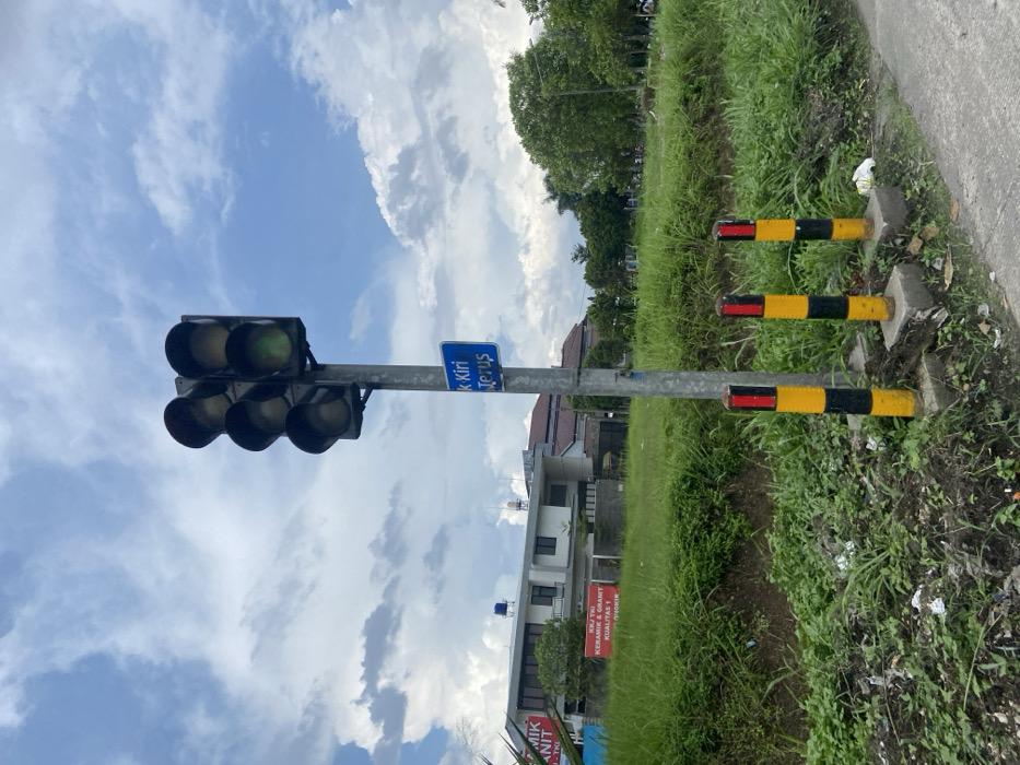 The image shows a traffic light with a green light, located on a street in Margaasih, Indonesia, at coordinates -6.97 latitude and 107.551 longitude. The traffic light is mounted on a gray metal pole, with three yellow and black striped posts positioned in front of it. A blue sign above the light says "K Kiri Terus," which translates to "Left Turn Straight." The pole is surrounded by lush green grass and bushes. In the background, a building with a red sign that says "KRIJ TRIO KERAMIK & GRANIT KUALITAS 1" can be seen. The sky is blue with white clouds.
