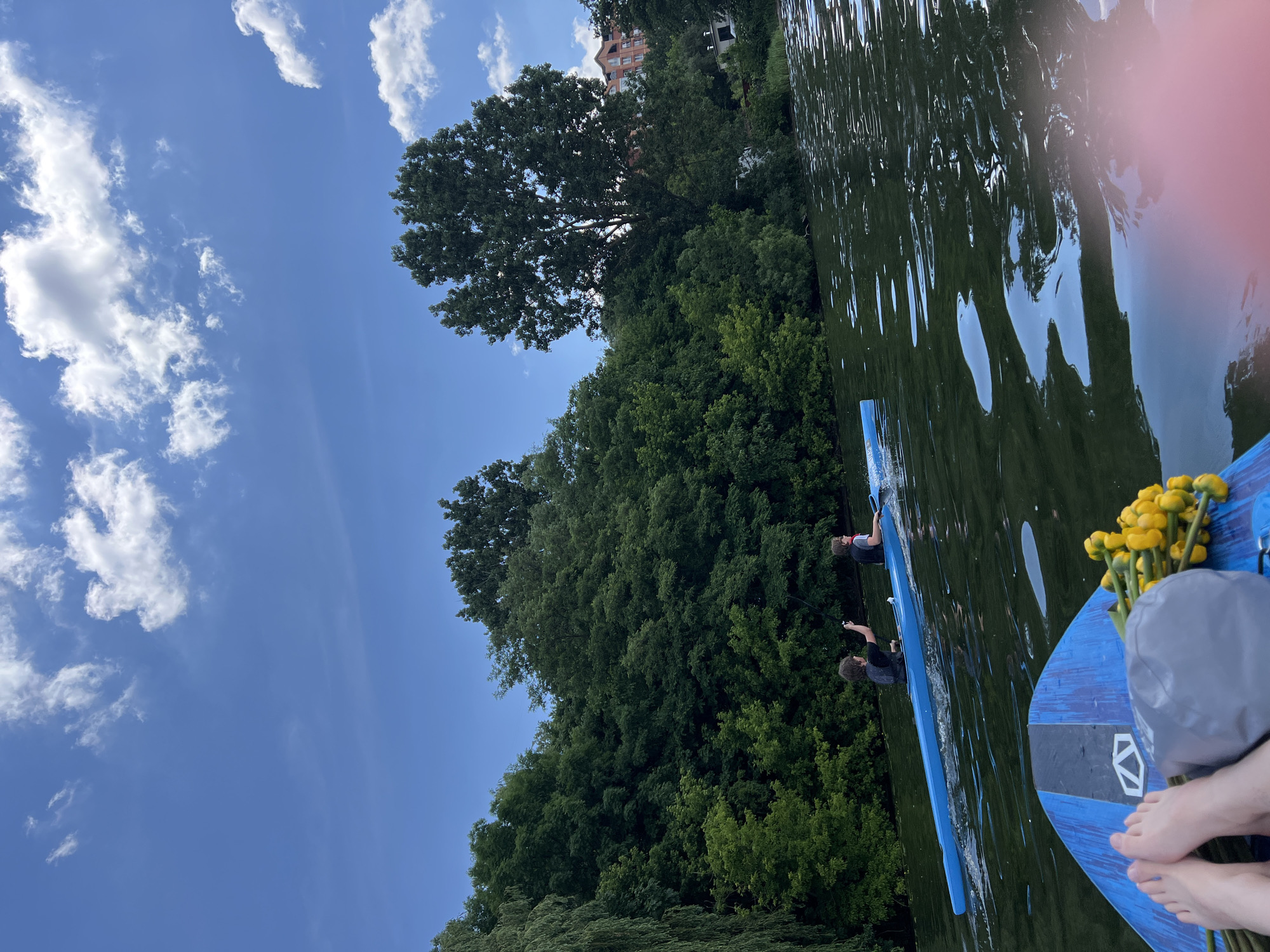 The image depicts a serene water scene with lush green trees lining the background. Two individuals are visible in a long blue kayak on the water, both wearing life vests. One person in the kayak is holding up a fishing rod. The foreground shows part of a blue paddleboard with a bouquet of yellow flowers and a gray bag. The sky is blue with scattered white clouds. The water is calm with gentle ripples. The overall setting suggests an outdoor recreational activity on a body of water, likely a lake or river, within the city of Ternopil, Ukraine, given the context.
