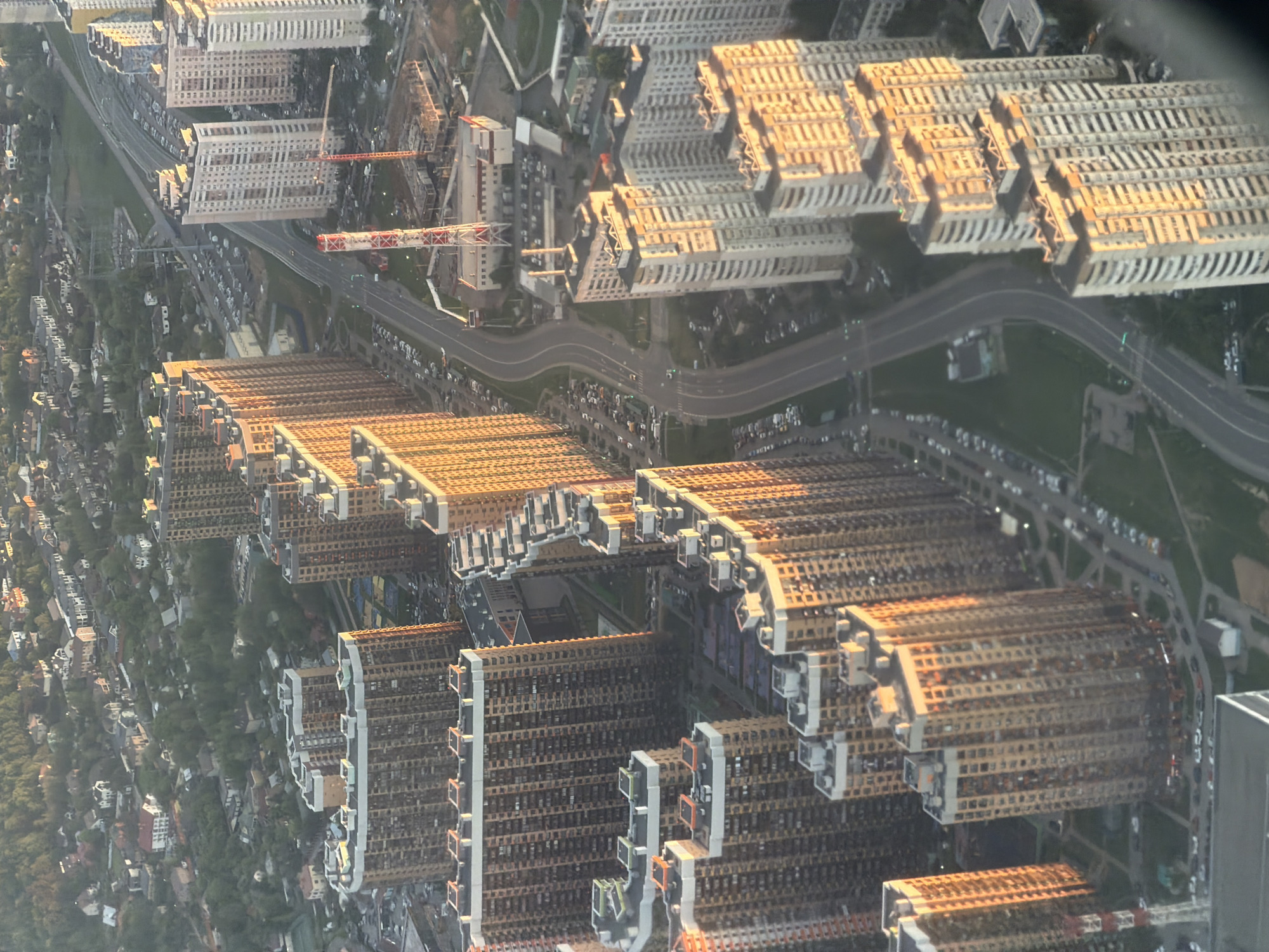 This aerial view depicts the "Solntsevo Park" district in Vnukovo, Russia. It shows dense clusters of high-rise residential buildings illuminated by sunlight, casting shadows on the structures. A winding road separates two clusters of buildings. Along the road, cars are parked, and some are in motion. In the upper portion of the frame, there's a construction site with a red and white construction crane. The rest of the surrounding environment consists of dense vegetation. The view implies the image was taken from an airplane.