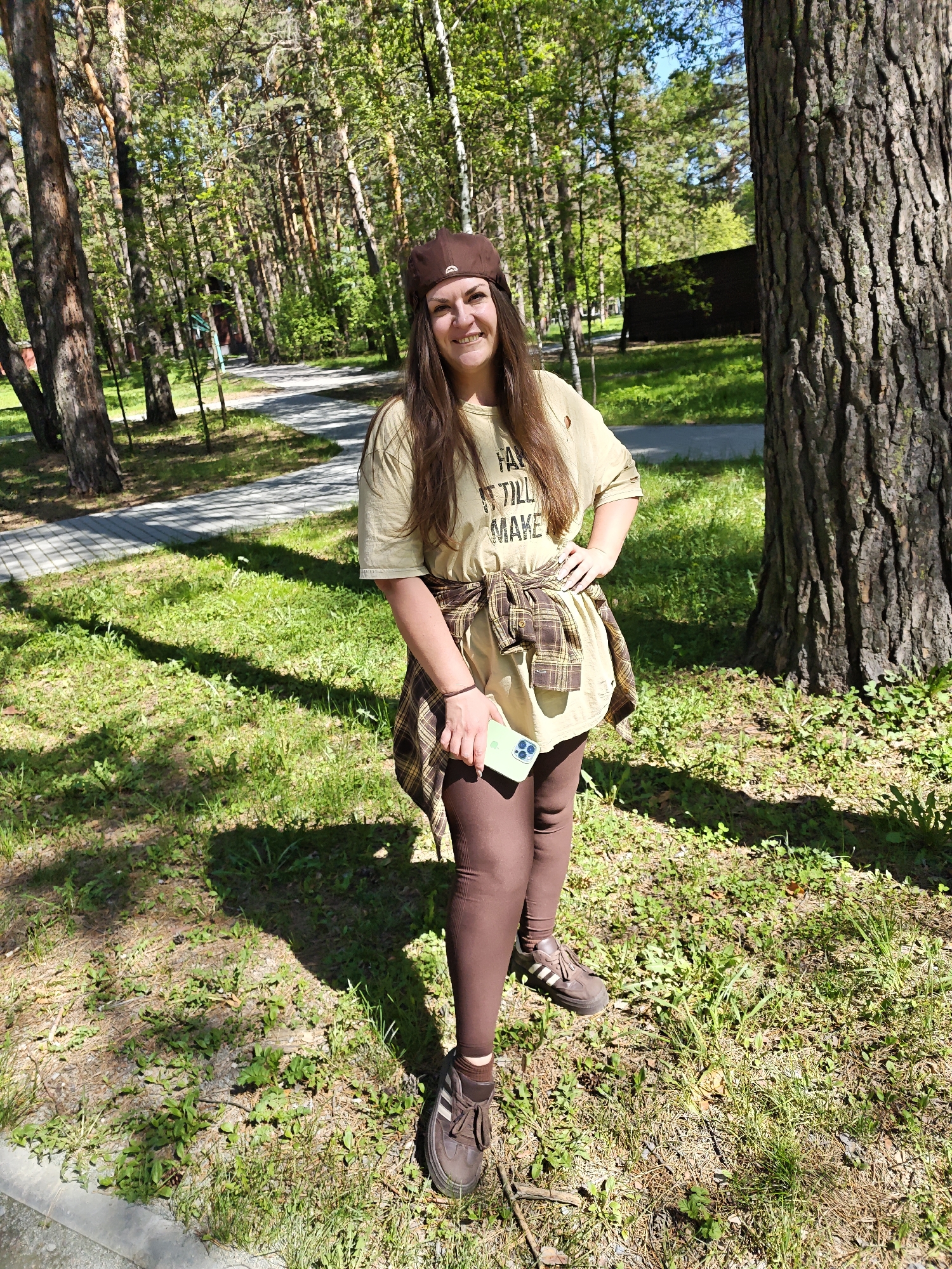 A young woman is standing in a grassy area, likely a park or wooded area, in Novosibirsk, Russia. She is smiling and facing the camera, wearing a brown hat, a tan t-shirt with black lettering, brown leggings, and brown sneakers. A plaid shirt is tied around her waist. She is holding a cell phone in her left hand. A large tree trunk is to her right, and other trees and a paved path are visible in the background. The lighting suggests it is daytime.
