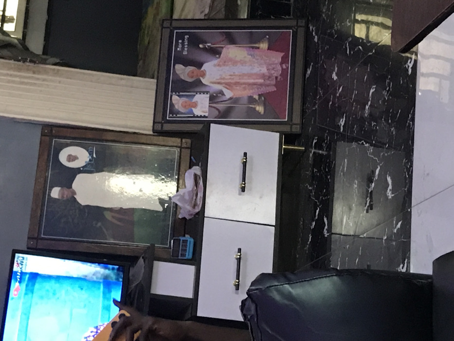 Two framed pictures are on a black and white marble wall above a white cabinet with two drawers. The top picture shows a person in an ornate outfit. The picture below shows a person in white attire. A small blue digital device sits on a shelf to the left of the cabinet. To the left is a television displaying a bright, colorful scene. A dark leather chair sits in the foreground.