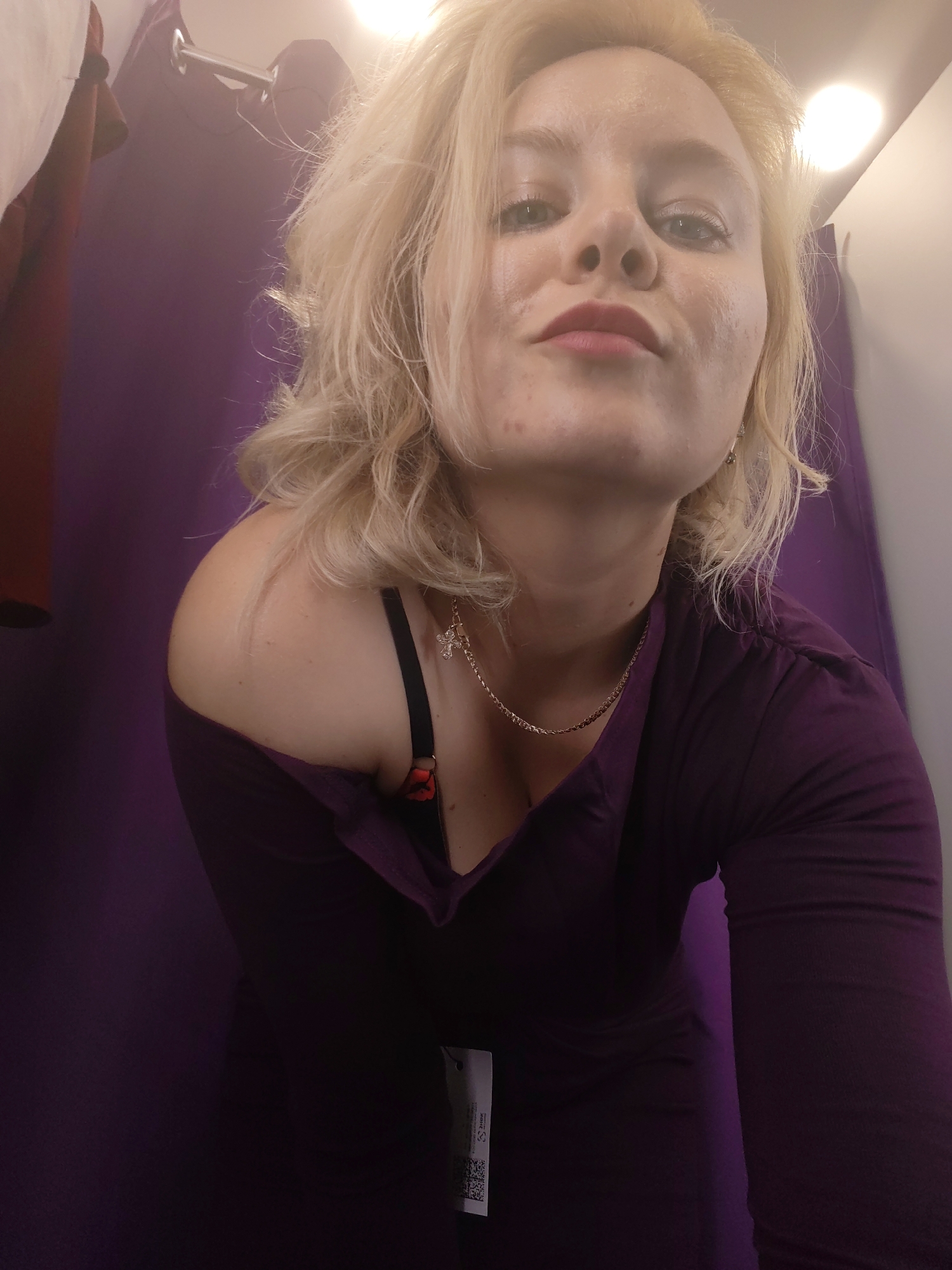 A young Caucasian woman with blonde, wavy hair is posing for a selfie in what appears to be a fitting room. She is wearing a purple, long-sleeved top that is off one shoulder, revealing a black bra with red accents. A thin gold chain with a cross pendant rests on her collarbone. The background consists of purple curtains and a bright overhead light. The woman has light skin with some blemishes and is looking up at the camera with her lips slightly parted. A white tag with a barcode is attached to her top. The scene is well-lit, suggesting an indoor setting, possibly a retail environment.