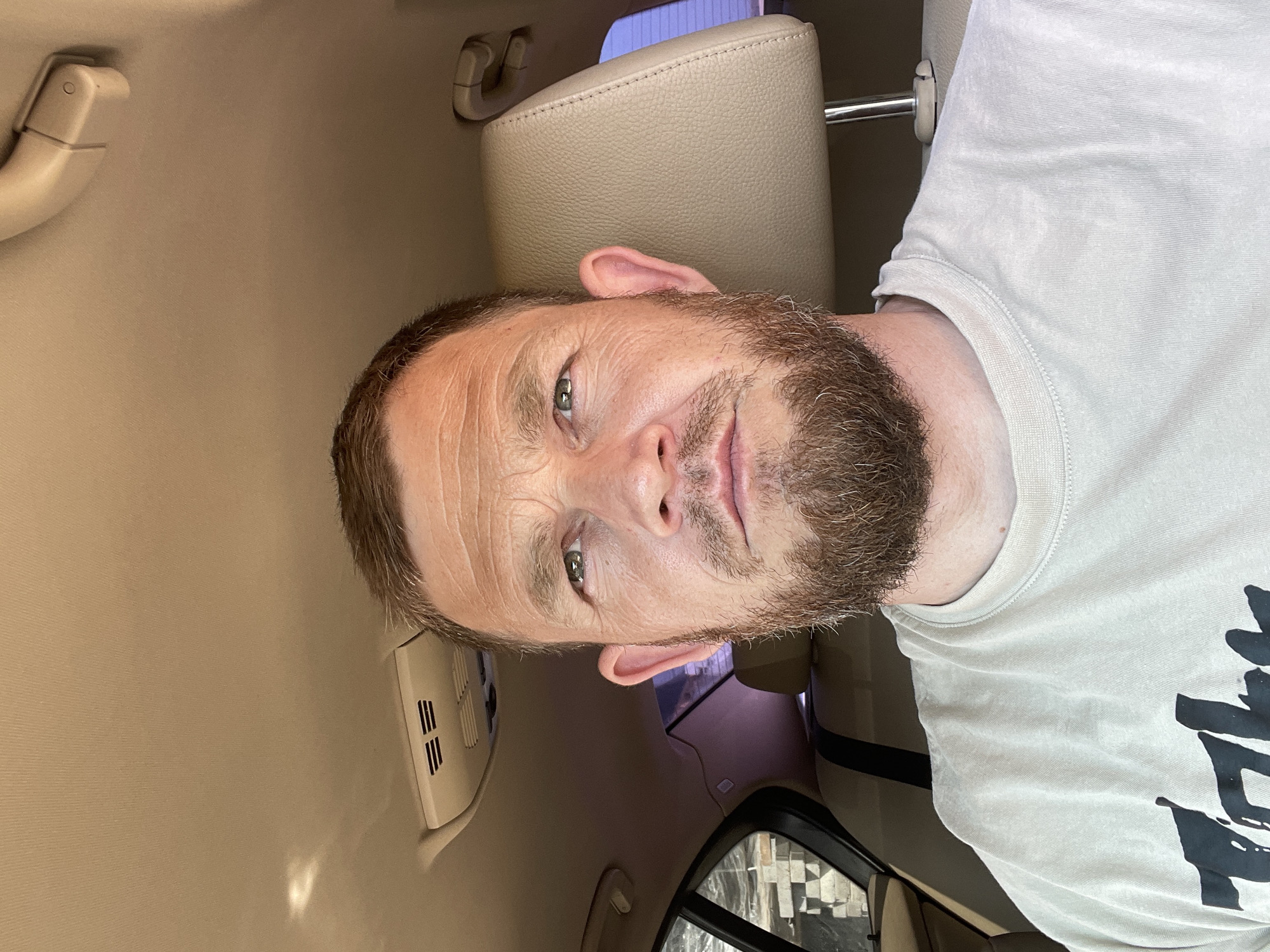 A man with a beard is in a car. The car's interior is beige. The car might be a sedan. The man is wearing a light-colored t-shirt. The image does not contain enough information to discern the location.