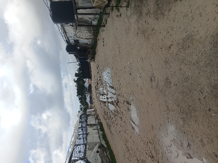The image depicts a dirt road traversing a semi-permanent settlement in the city of Khan Yunis, Palestinian Territory, under an overcast sky. The unpaved road, seen from a low perspective, is significantly muddy and contains numerous puddles, suggesting recent rain or poor drainage. To the left, a large structure with a metal frame and torn white sheeting or tarpaulins appears partially collapsed or damaged. To the right, a row of informal structures or shelters with white-washed walls or covered with white tarps extends into the distance. Elevated black cylindrical water tanks are regularly spaced along the right side, mounted on metal or wooden stands next to these structures. Distant trees and residential buildings are visible on the horizon beneath the cloudy sky. No individuals, vehicles, or specific actions are discernible within the frame.