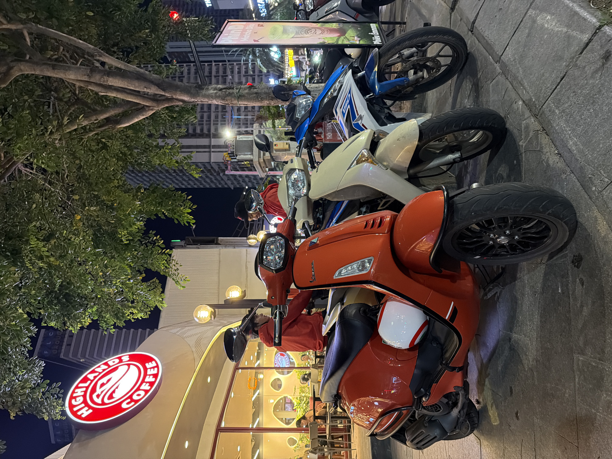 A nighttime scene in South Quy Nhon Ward, Vietnam, features several parked scooters and motorcycles lined up along a sidewalk. The foreground is dominated by a bright orange Vespa scooter, with a white helmet adorned with red and green stripes resting on its seat. Behind it, a beige scooter and a blue and white motorcycle are also parked.
The scooters are positioned in front of a building with a large, illuminated sign that reads "HIGHLAND COFFEE" in a red circular logo. The building's facade has large windows, revealing a warmly lit interior with patrons inside. Outdoor lighting fixtures, resembling spherical lamps, hang above the coffee shop entrance, casting a soft glow on the scene.
A large tree with leafy branches extends from the left side of the frame, its trunk and limbs providing a natural element to the urban setting. In the background, tall buildings and streetlights indicate a bustling city environment. The overall impression is one of a common urban scene at night, likely a busy area with a popular coffee shop. The image is captured in portrait orientation.