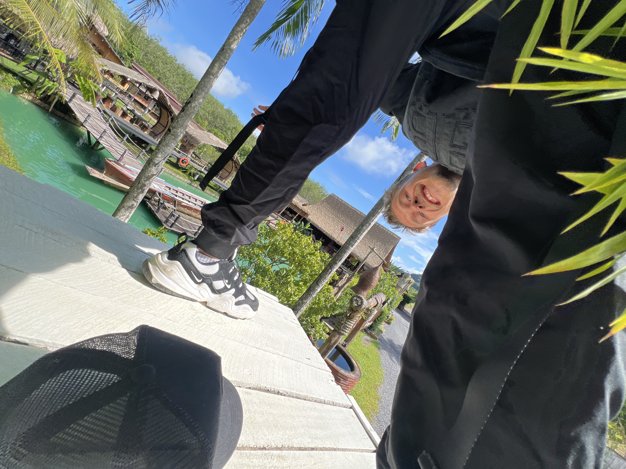 This is an upside-down, low-angle selfie taken outdoors. The main subject is a young man with blonde hair, blue eyes, and a wide smile, looking directly at the camera. He is wearing black pants and a black denim vest over a black shirt. His left leg is bent, with a white and black athletic shoe visible. A black baseball cap with a mesh back is lying on a light-colored wooden surface in the foreground.
The background features a vibrant green body of water with wooden walkways and small boats. Several thatched-roof structures are visible along the water's edge, surrounded by lush green trees and palm trees. In the distance, there are more trees and what appears to be a road. The sky is bright blue with scattered white clouds, suggesting a sunny day.
The scene is set in a tropical or subtropical location, with the water, vegetation, and architectural style hinting at a resort or a natural park. The overall mood is cheerful and relaxed, conveyed by the man's enthusiastic smile. The time of day appears to be daytime, likely late morning or early afternoon due to the bright sunlight and clear sky. No text is visible in the image. The location context provided is Tambon Pa Kae Bo Hin, Thailand.